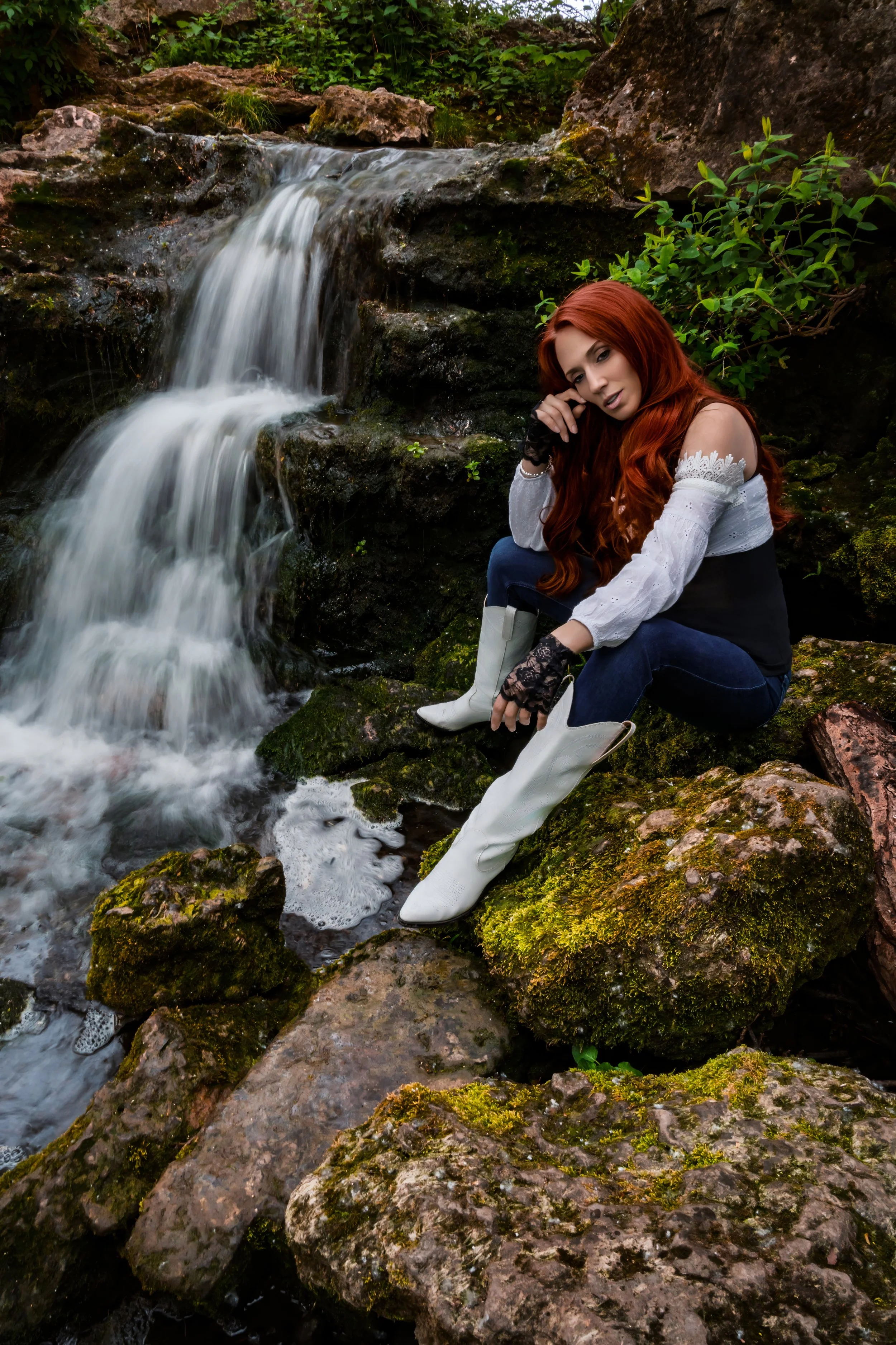 Stylised portrait of a red haired woman wearing vintage white corset,  white boots, blue jeans and lace gloves by a waterfall in Cheltenham 