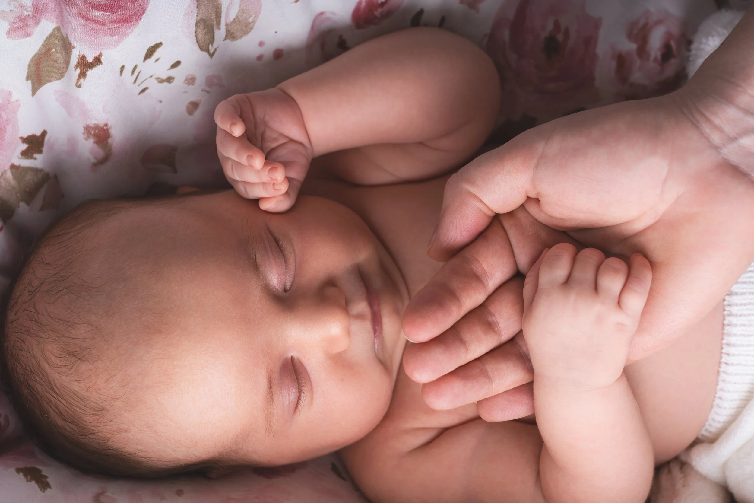 A sleeping baby holds her mother's hand on a floral-patterned blanket. The scene conveys tenderness and warmth, with soft lighting enhancing the gentle mood.