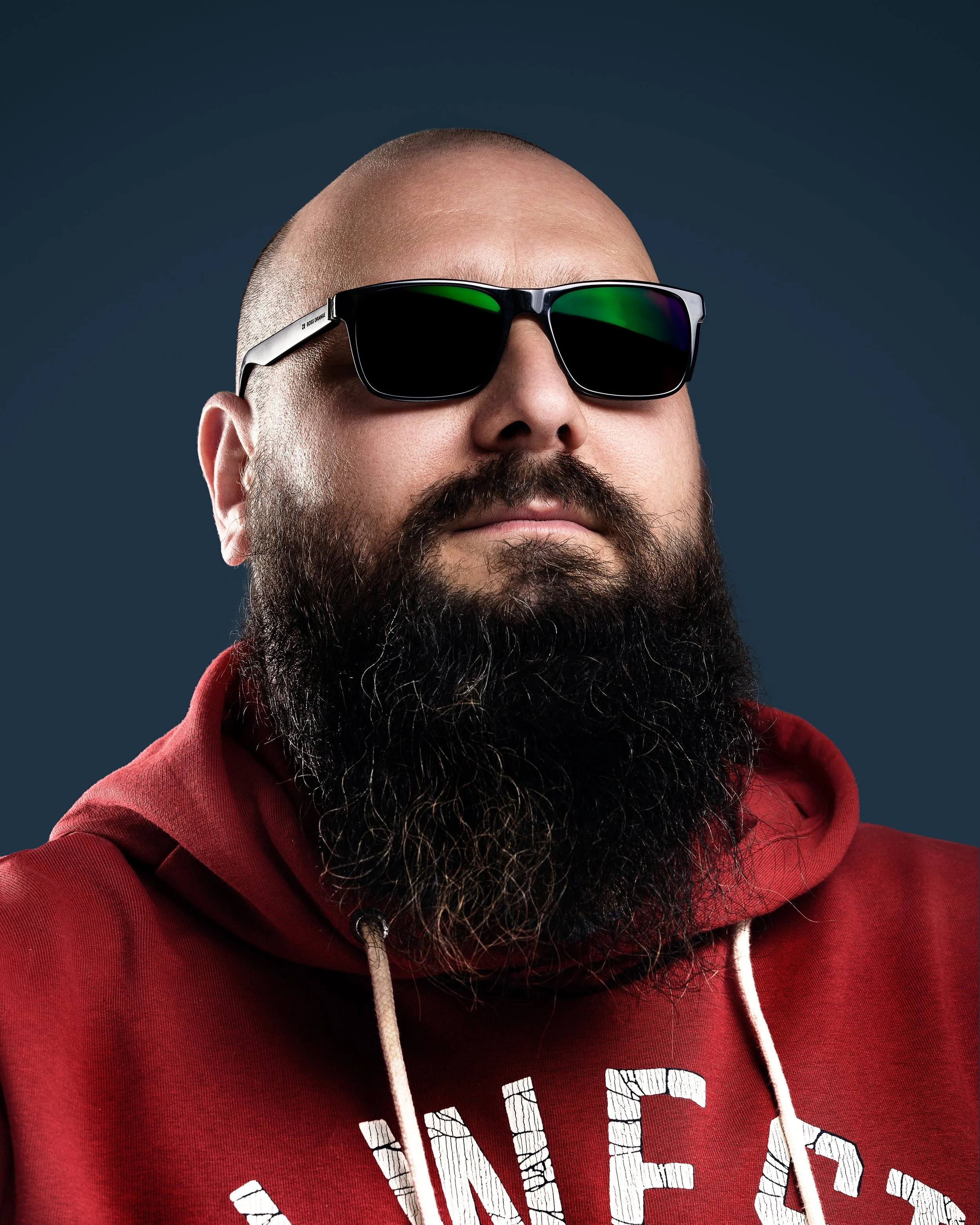 Portrait of a bald man with a thick beard wearing sunglasses and a red hoodie. The background is is a dark blue gradient conveying a confident mood