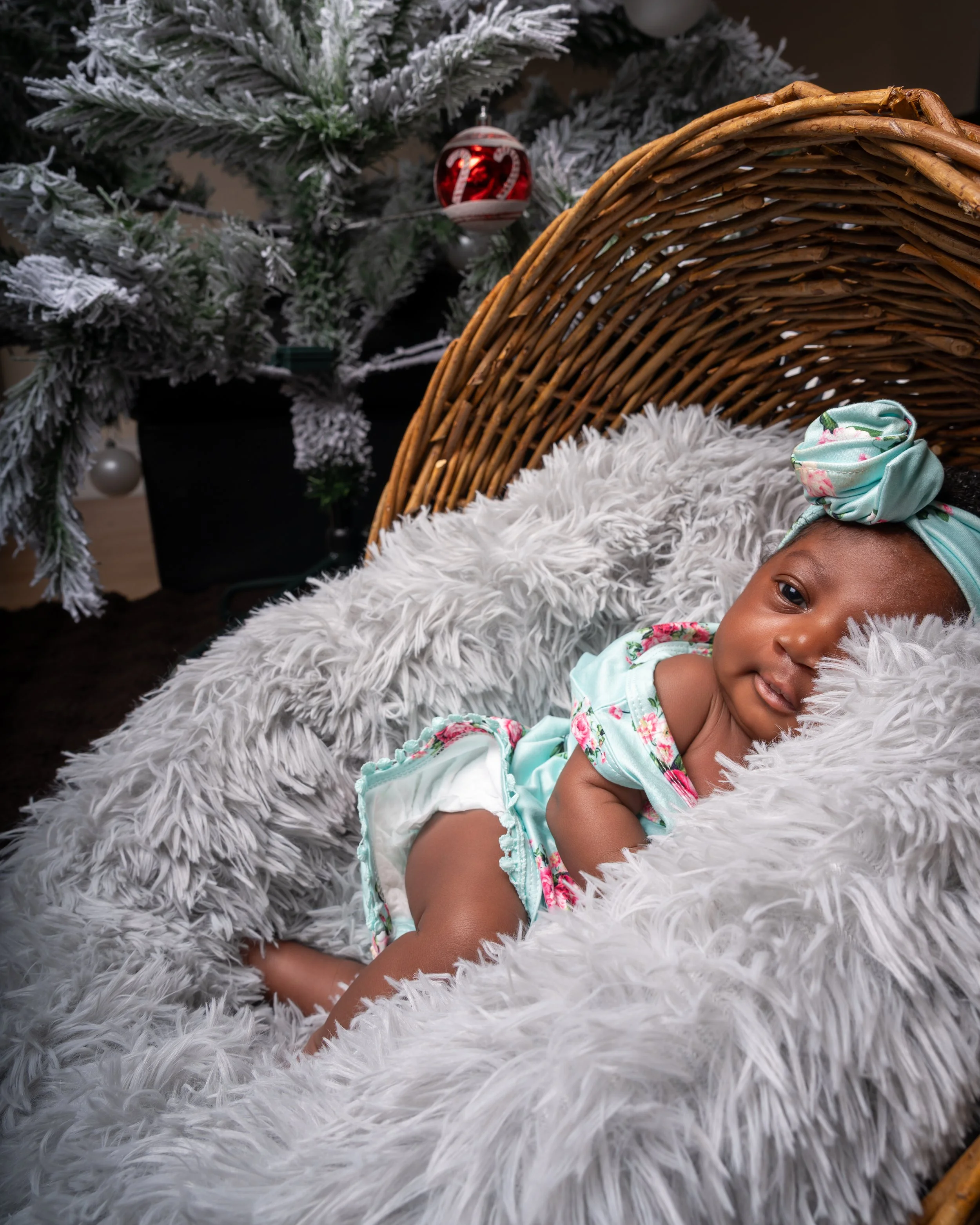 A baby wearing a mint green outfit and headband lies in a fluffy, gray-lined basket. A frosted Christmas tree with ornaments is in the background, adding a festive touch.
