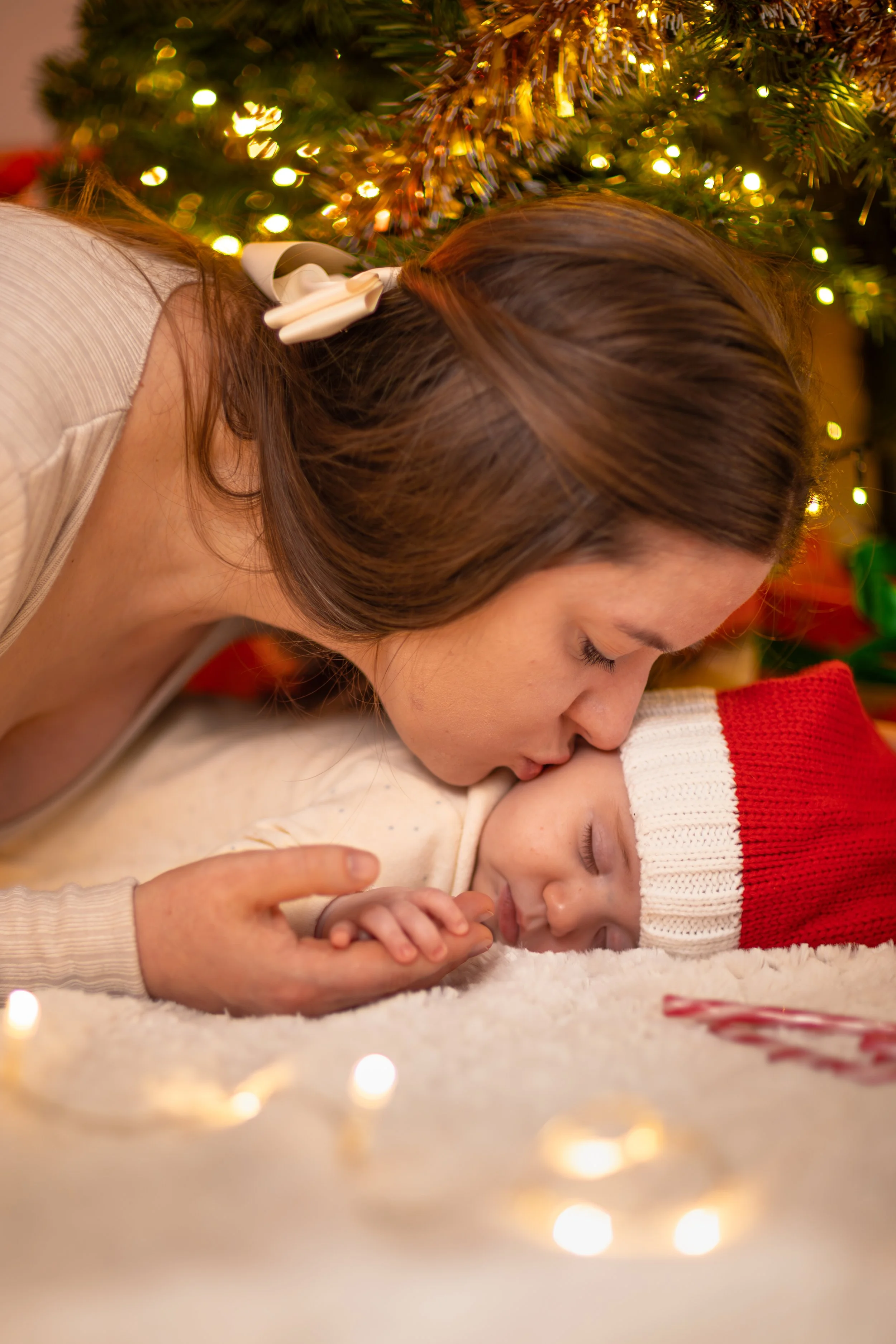 A woman lovingly kisses her baby's forehead under a Christmas tree adorned with lights. The baby, in a red Santa hat, sleeps peacefully. Warm, festive ambiance.