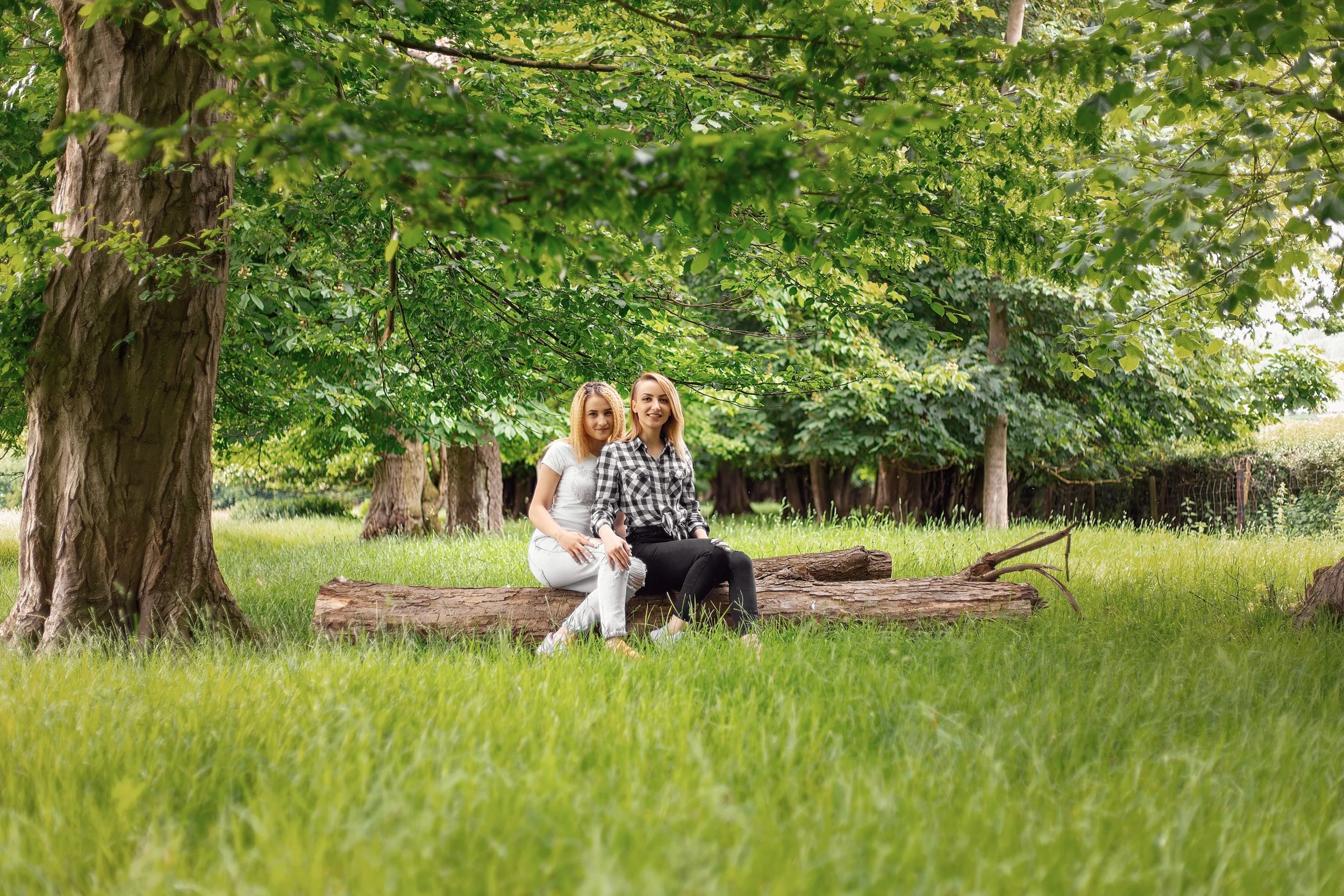 Two blond young women are sitting on a tree trunk in the woods