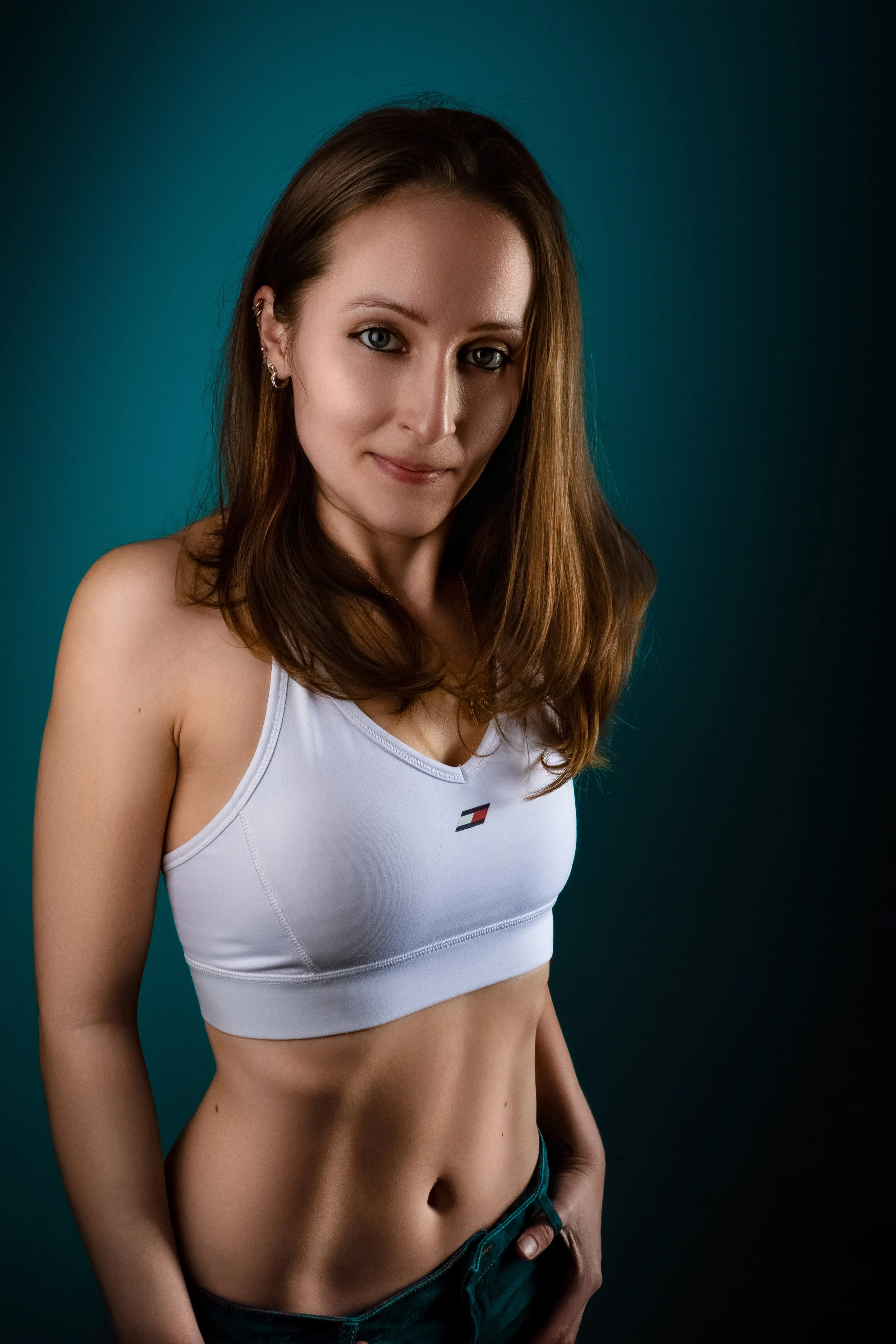 Dramatic portrait of a young woman with the soft side light which accentuates her tones. The blue gradient background compliments her skin. She's wearing white crop top and blue jeans. Smiling and standing with Confidence