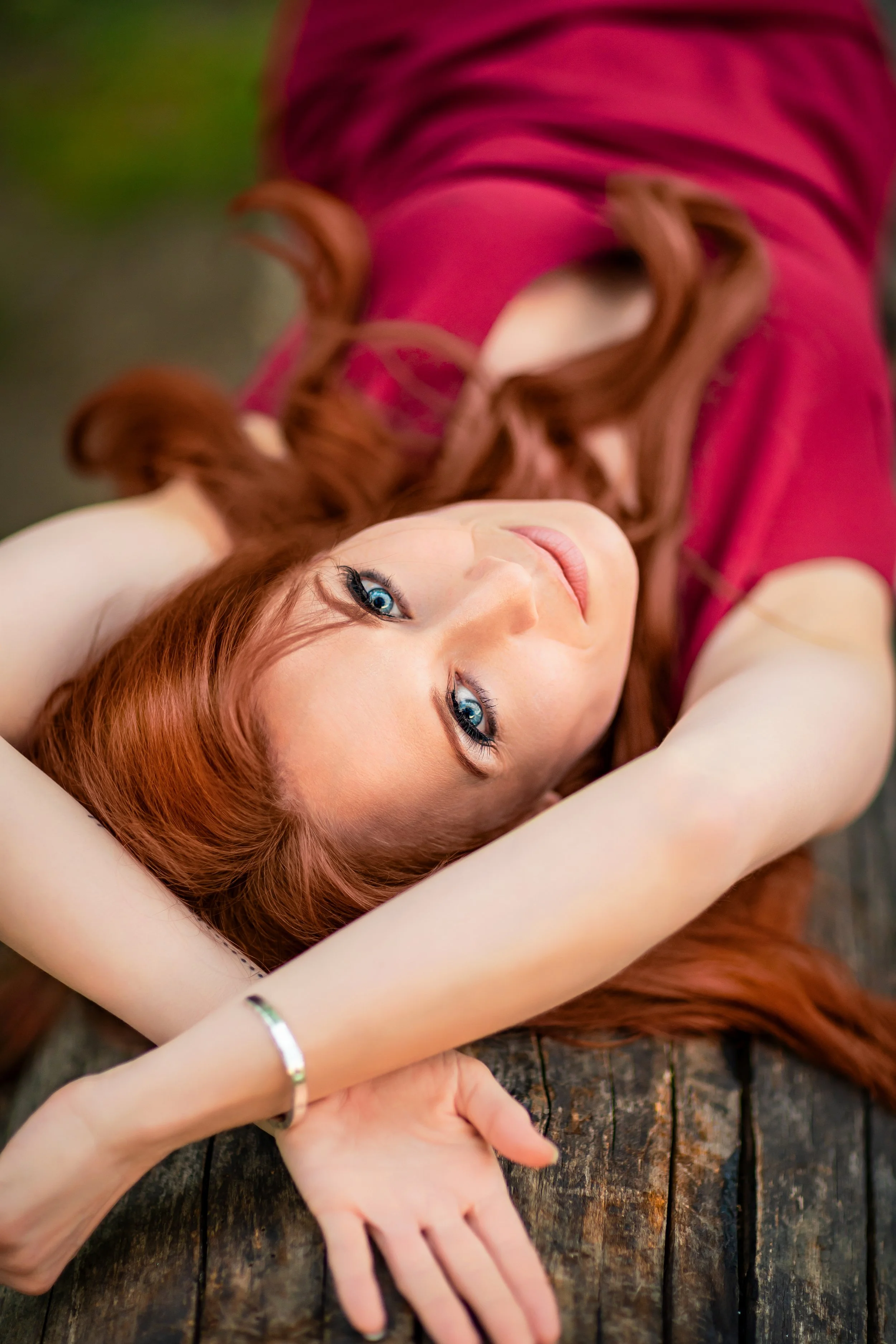 Portrait of a woman with long red hair is lying on her back on a wood trunk, wearing a red top. She gazes upward to the camera with a calm expression, arms extended and crossed overhead 