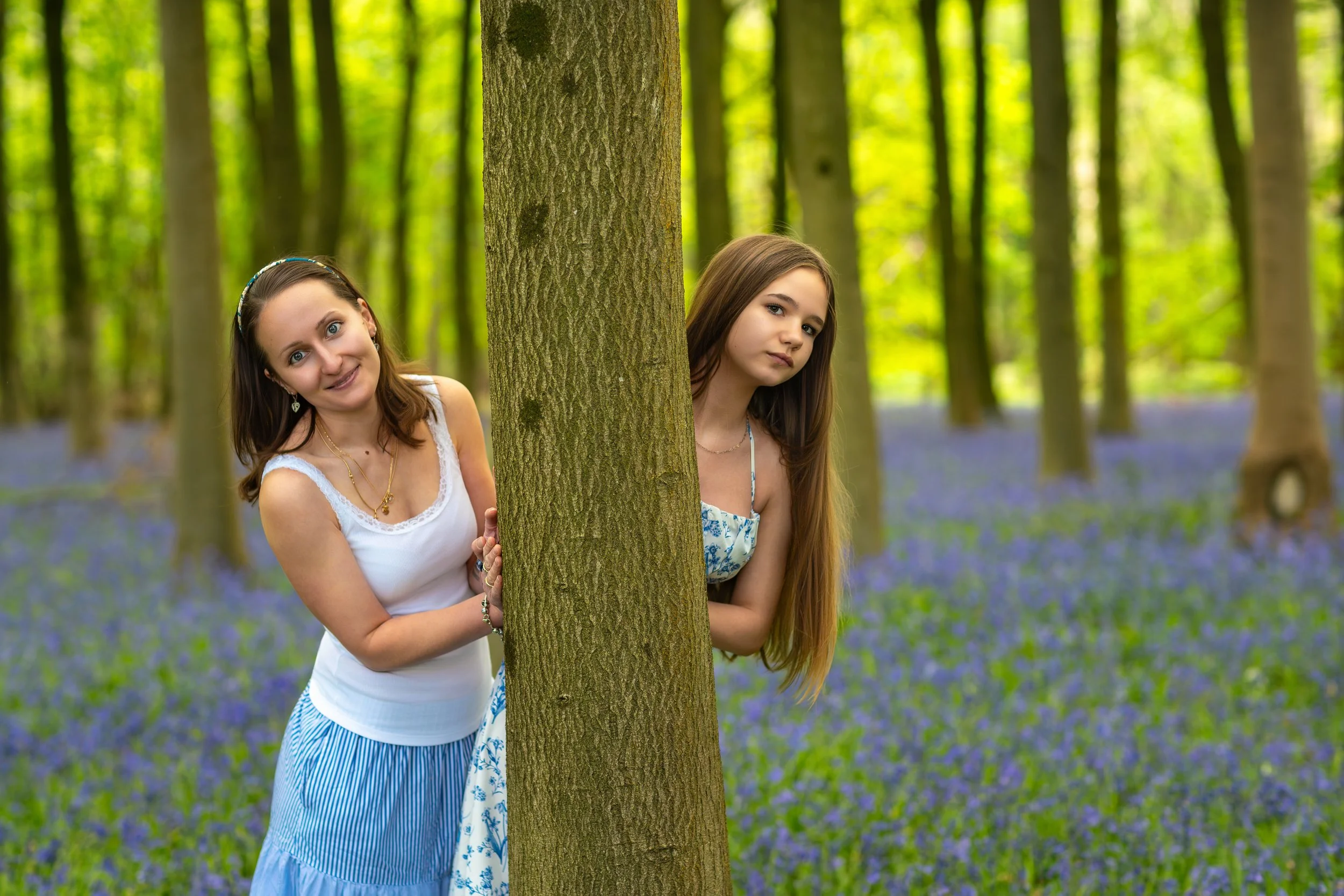 A woman and her niece playfully peeking from behind a tree in a woodland carpeted with blooming bluebells and soft spring light.