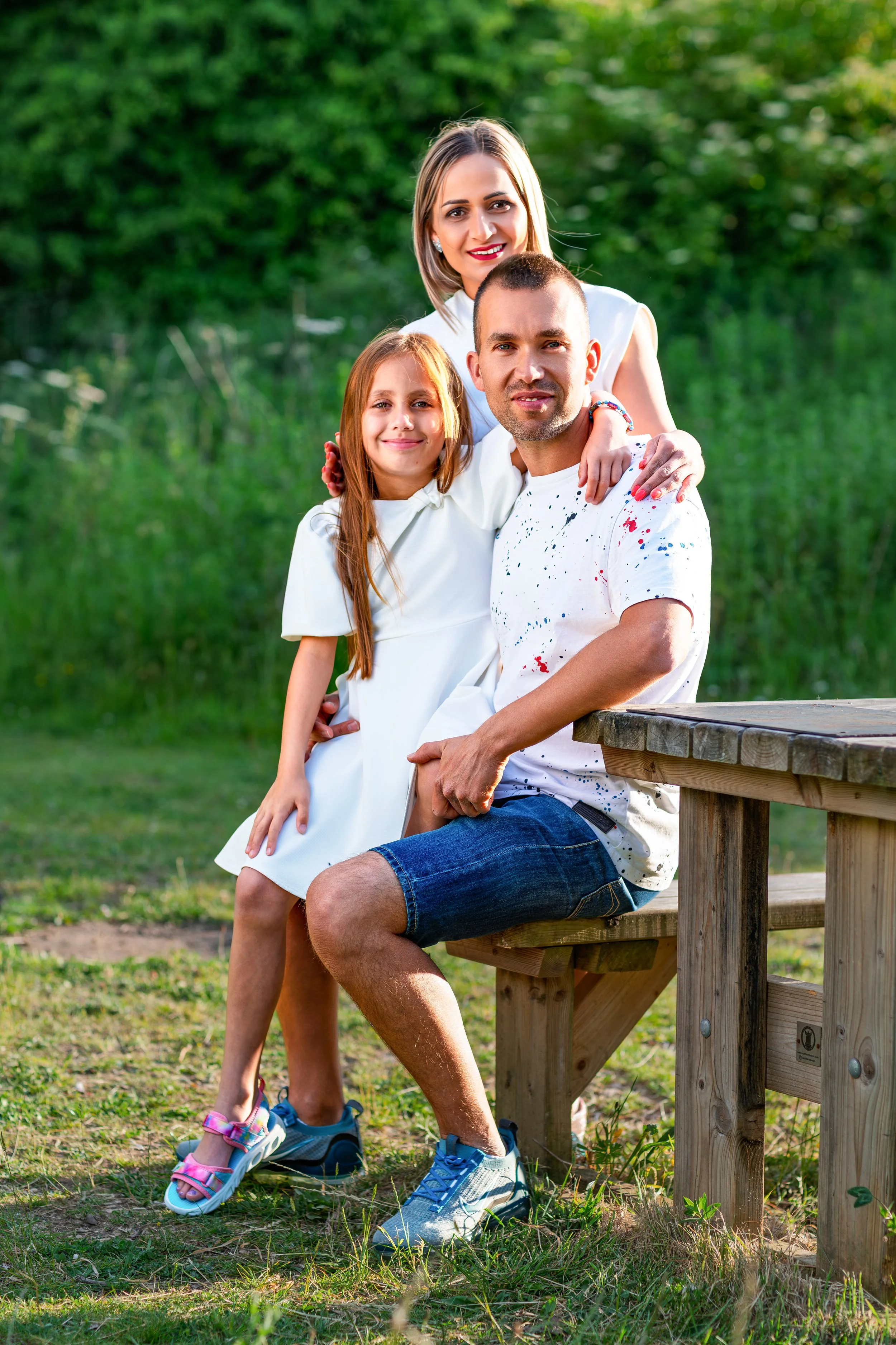 A family of three poses in a sunny park. A girl in a white dress sits on her father's lap, who wears a white shirt and denim shorts. The mother stands behind them, smiling.