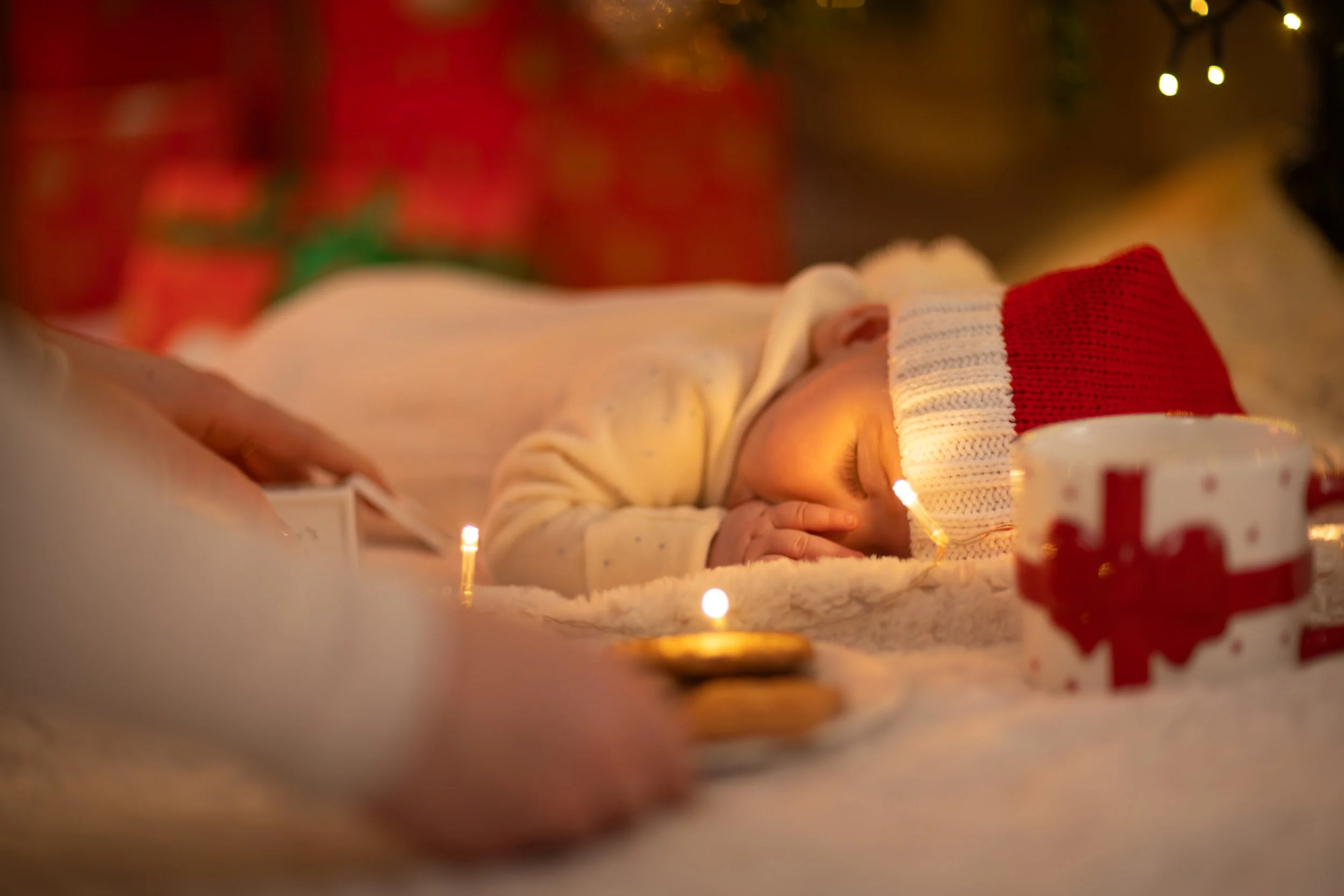 A sleeping baby in a Santa hat lies on a soft blanket surrounded by Christmas lights, and wrapped gifts. A hand places cookies nearby, creating a warm, festive atmosphere.