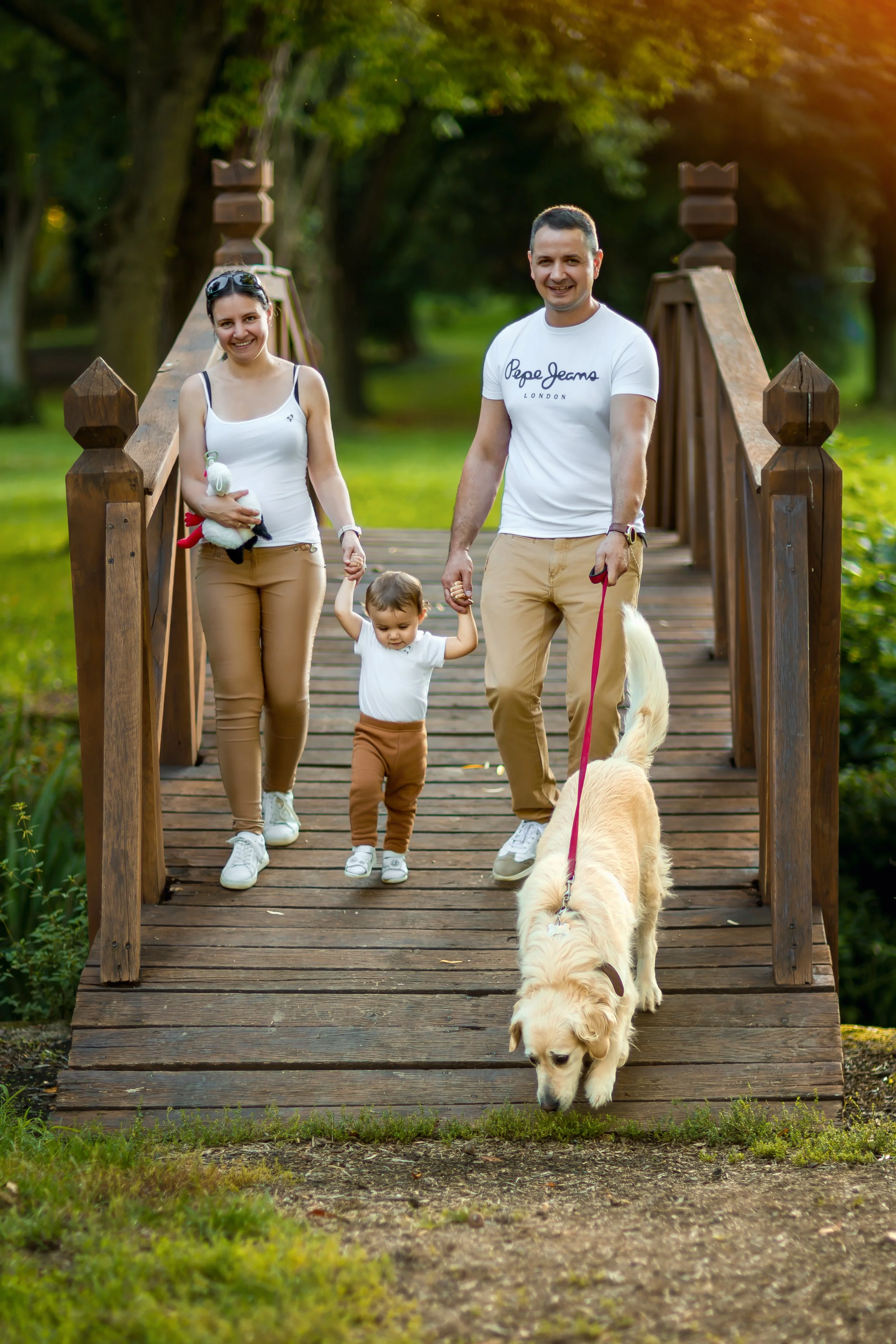 A happy family walks on a wooden bridge in a park, holding hands. The father holds a dog on a leash. The setting is sunny and peaceful.