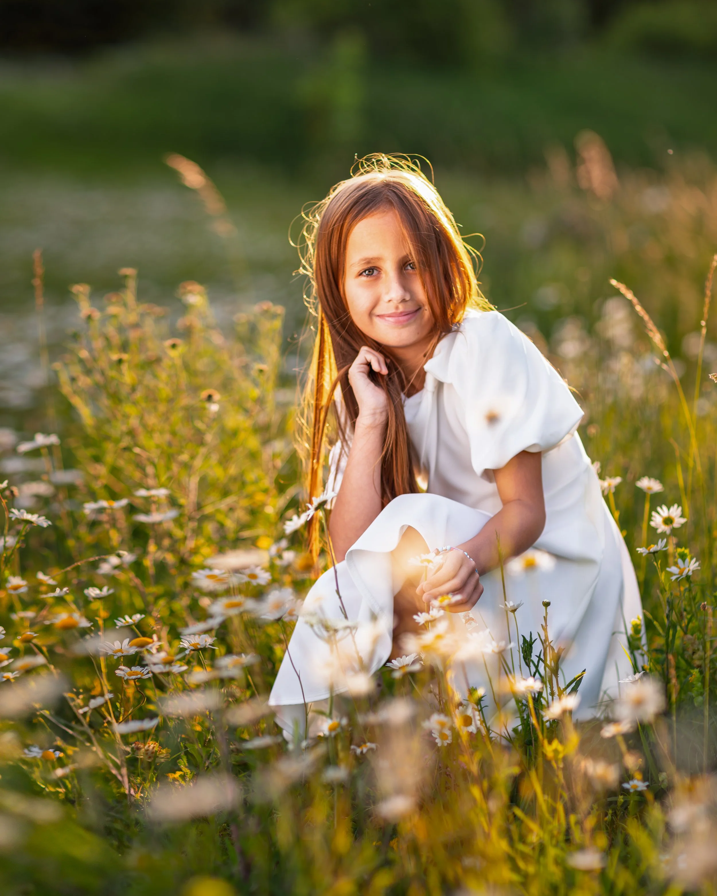 Portrait of a young girl in a white dress sits in a sunlit park, surrounded by wildflowers. She smiles gently, her hair glowing golden in the soft evening light.