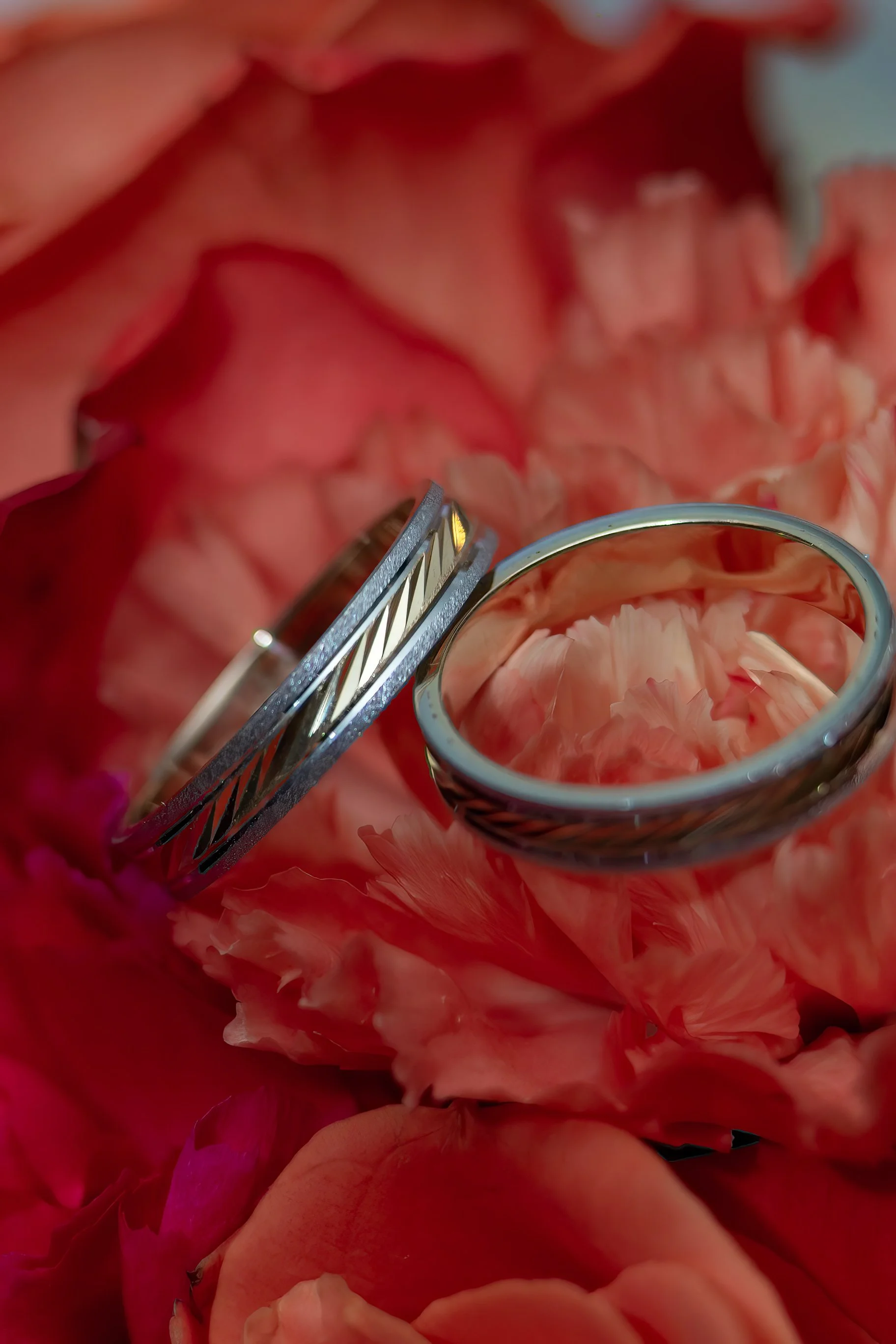 Wedding bands with engraved patterns rest on vibrant pink and red flowers. The image conveys a romantic and elegant tone, highlighting love and commitment.