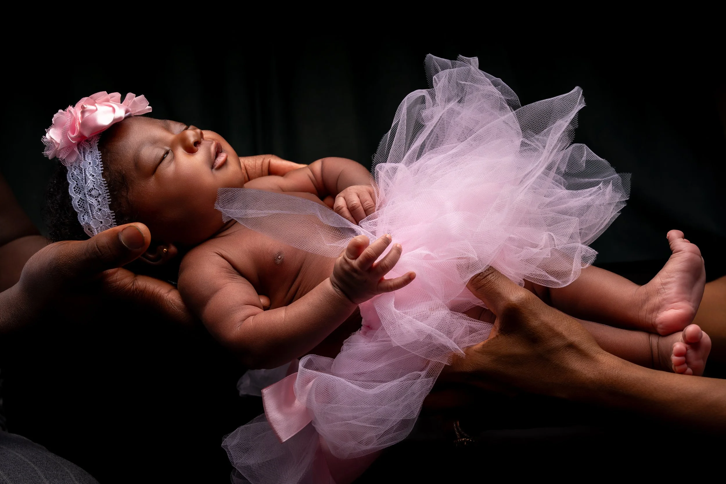 A peaceful newborn in a pink tutu and floral headband is gently cradled by two adult hands against a dark background, conveying warmth and tenderness.