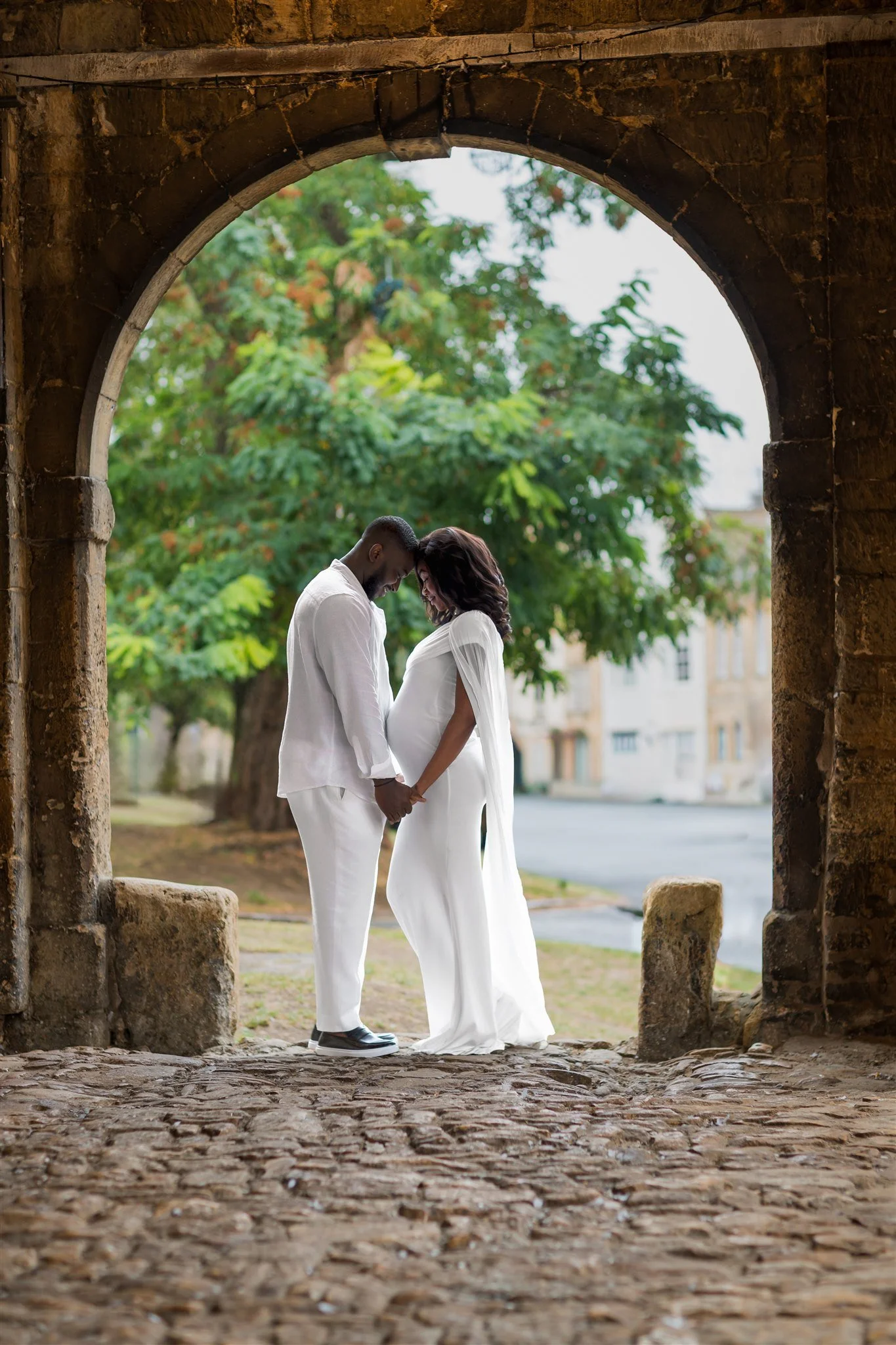 A man and a pregnant woman dressed in white stand under stone arch, foreheads touching, holding hands. A tree and street are visible in the background, conveying tranquility and love.