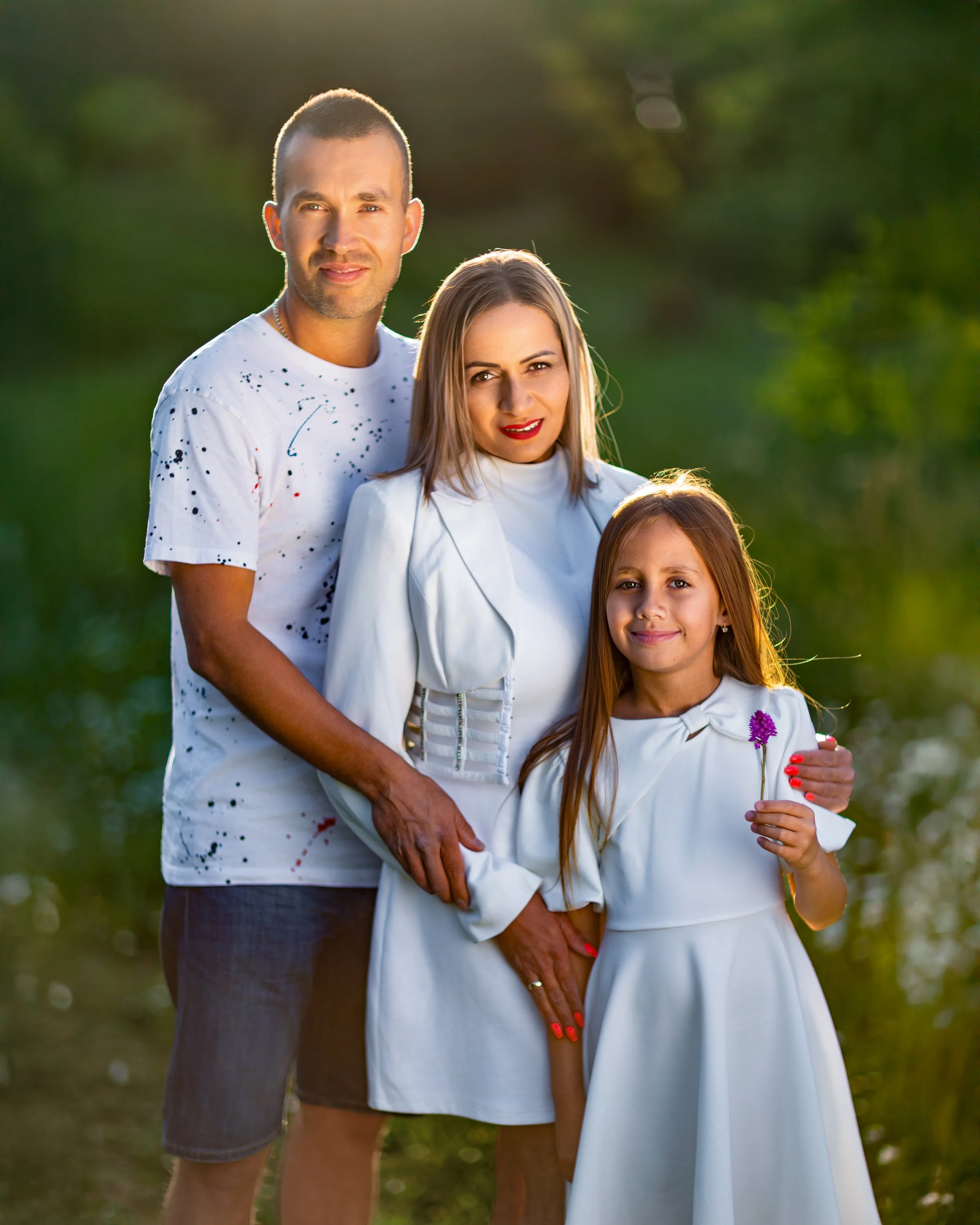 Portrait of a family of three poses outdoors, smiling softly. The man, in a white shirt, stands beside the woman and girl, both in white dresses, against a lush, green background.