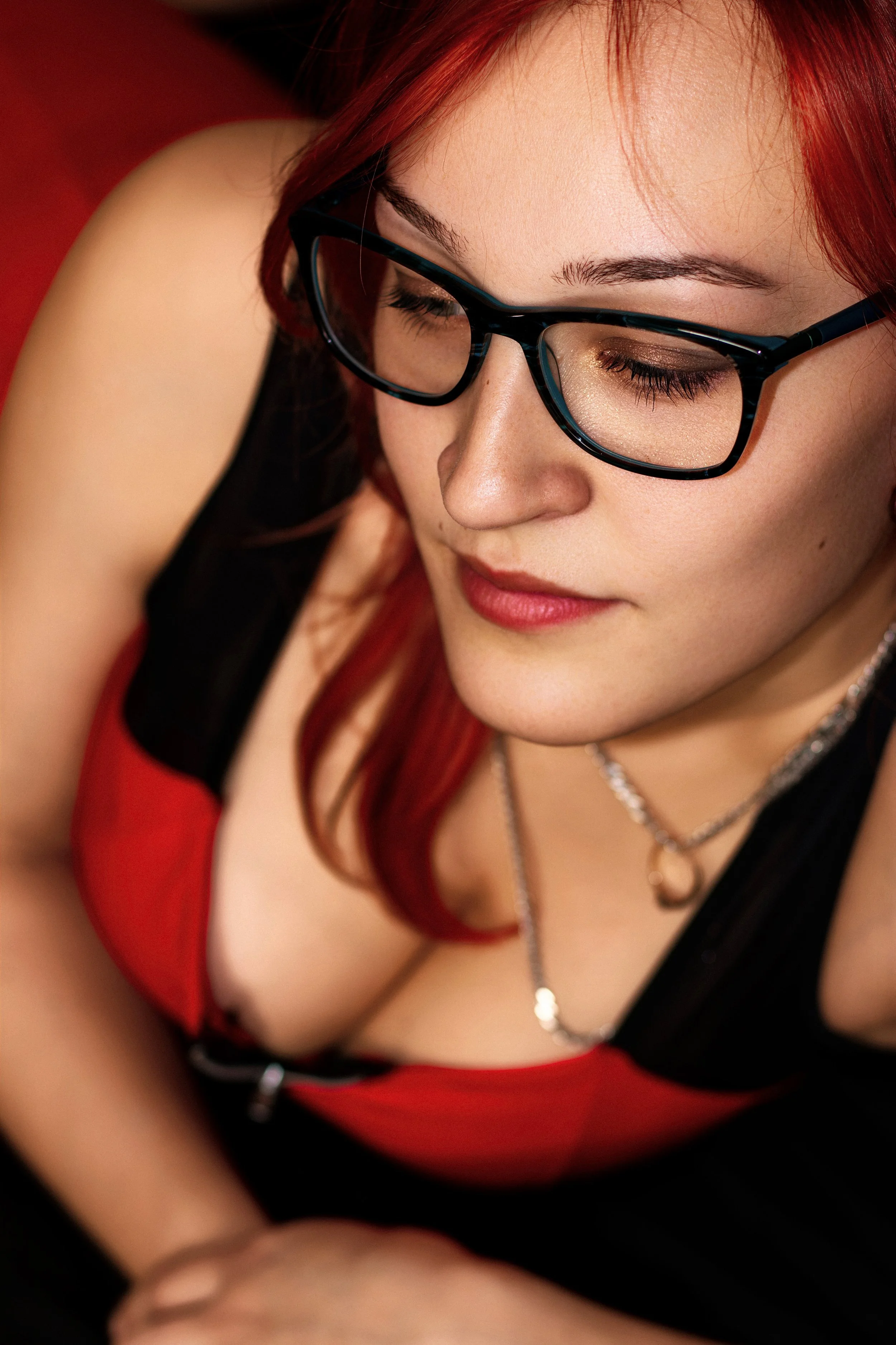 A woman with red hair and glasses gazes downwards, wearing a black and red top. The lighting is warm, and her expression is calm and thoughtful.