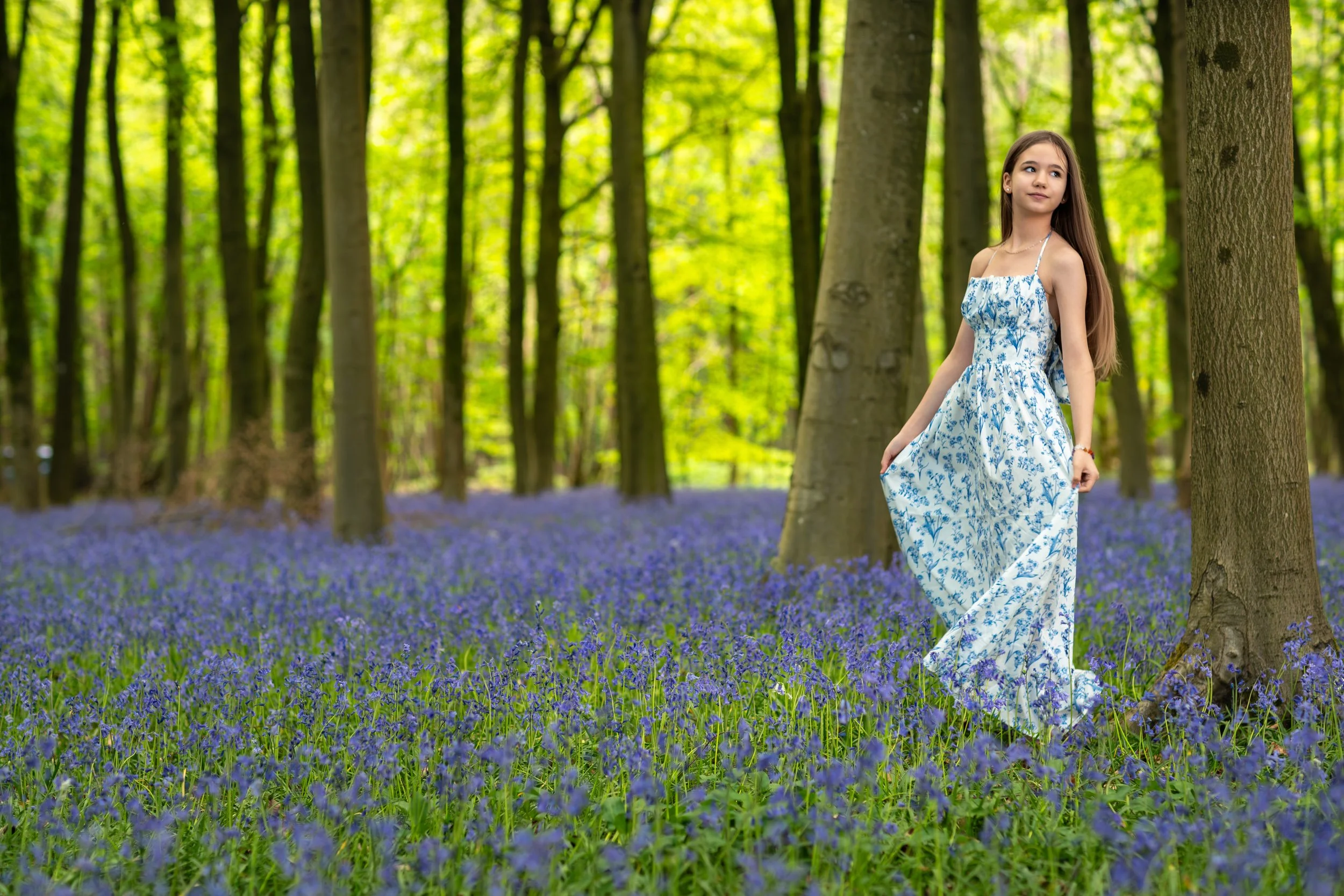 A young girl standing gracefully among blooming bluebells in a sunlit woodland, wearing a flowing floral dress and surrounded by vibrant spring colours.
