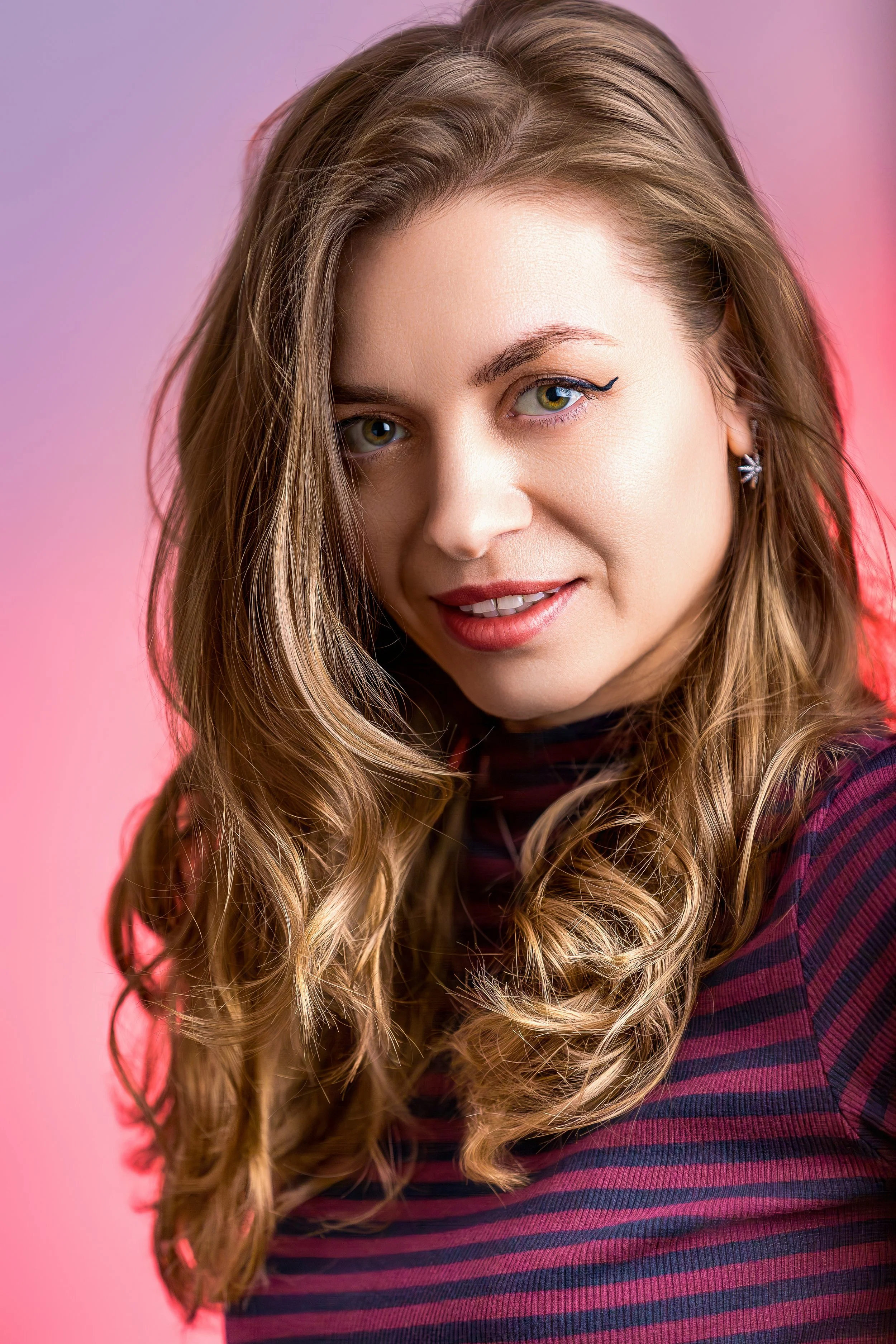 Portrait of a woman with long wavy hair smiles subtly against a pink and purple background. She wears a striped top and a subtle makeup, conveying a warm and friendly vibe