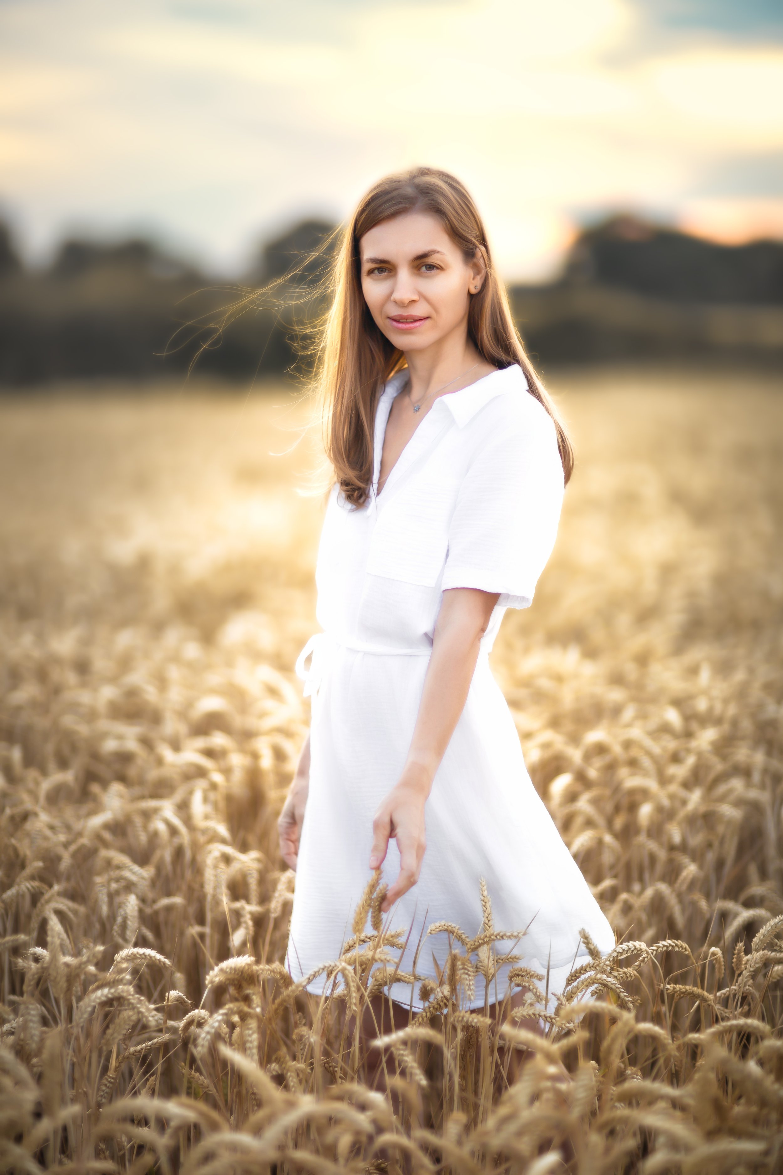 Melancholic portrait of a woman in the sunset on a wheat field wearing white summer dress 