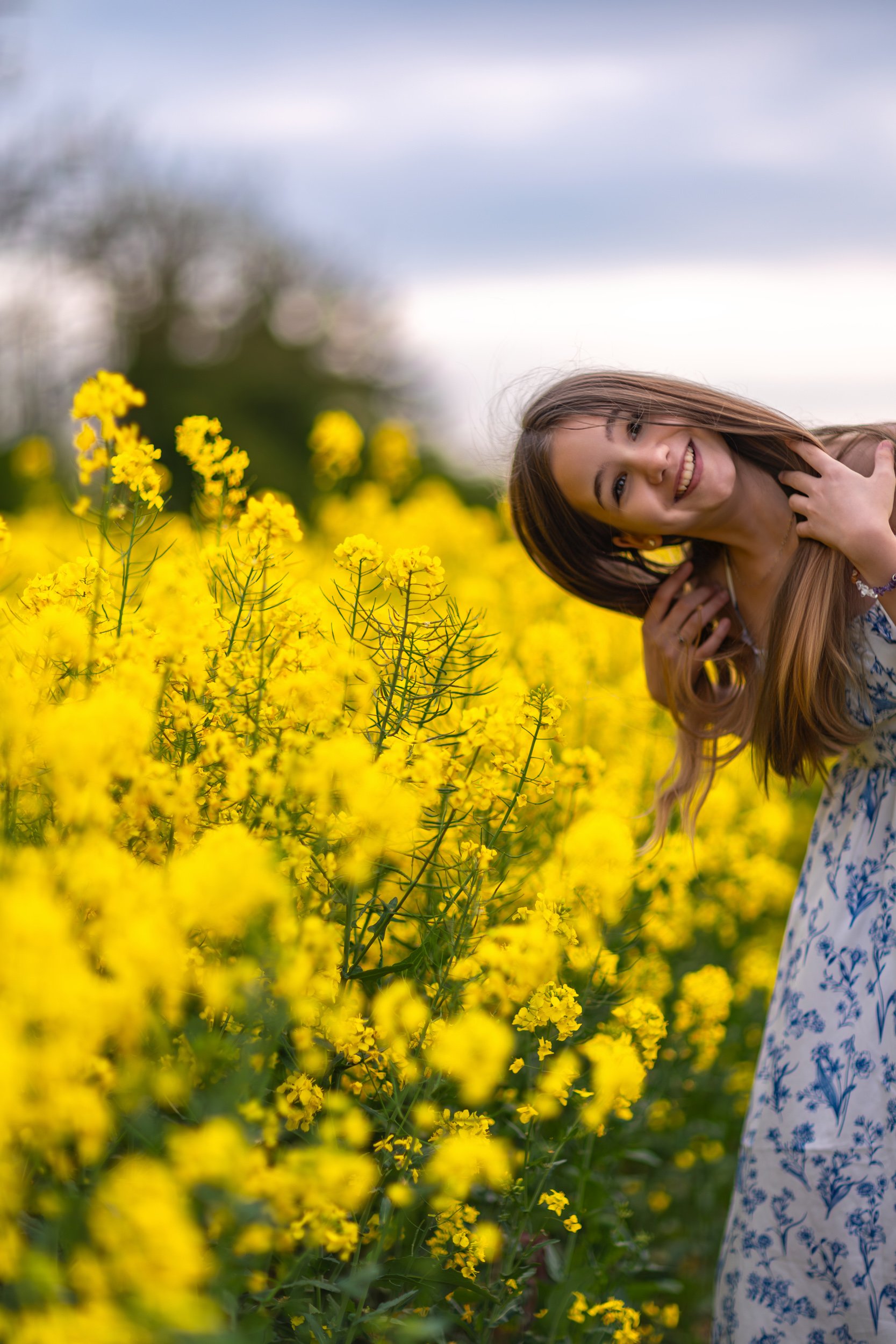 A smiling young girl leaning into a field of blooming yellow rapeseed flowers, enjoying a bright and playful spring evening outdoors.