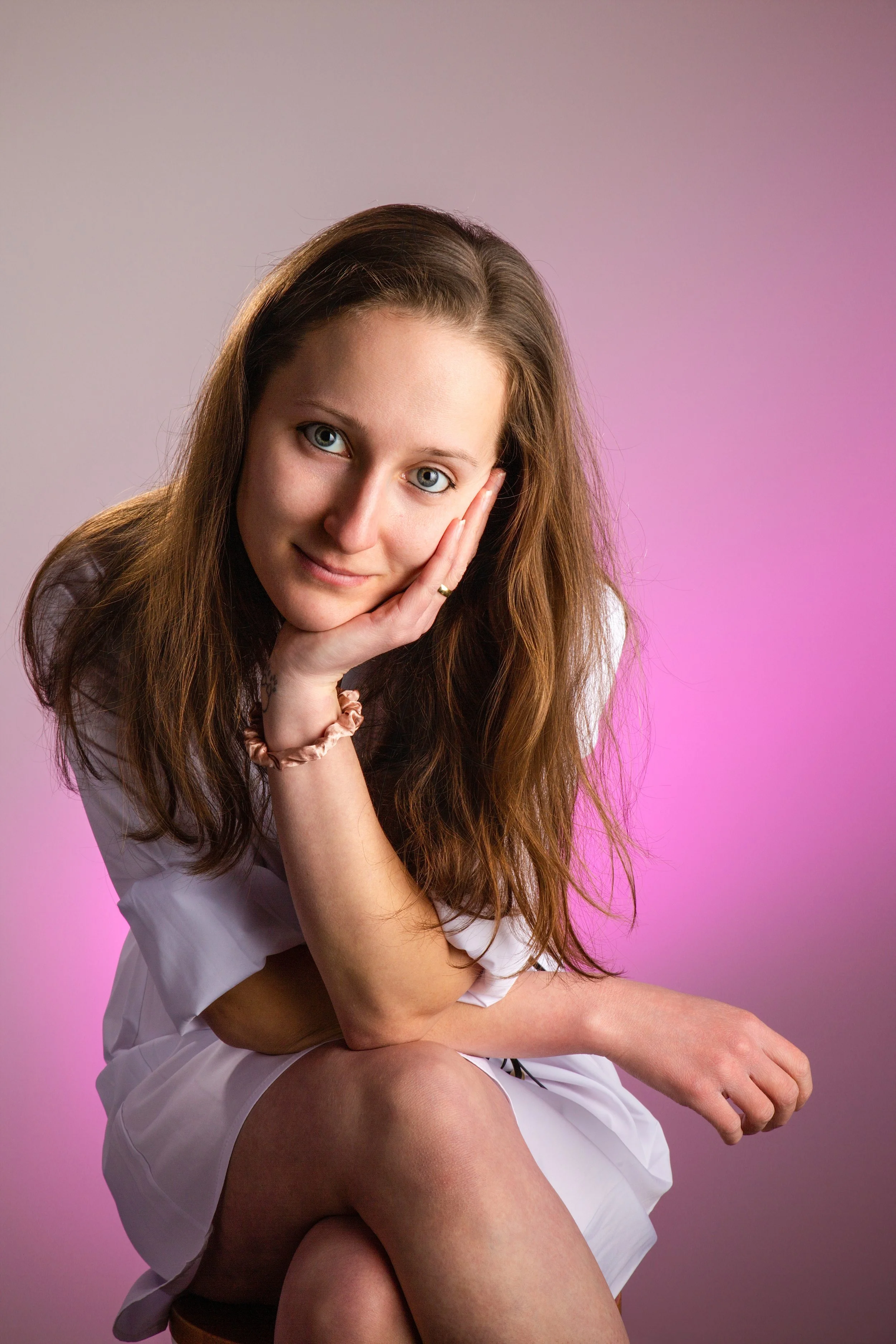 Indoor portrait of a young woman with brown hair in a white dress against a pink background 
