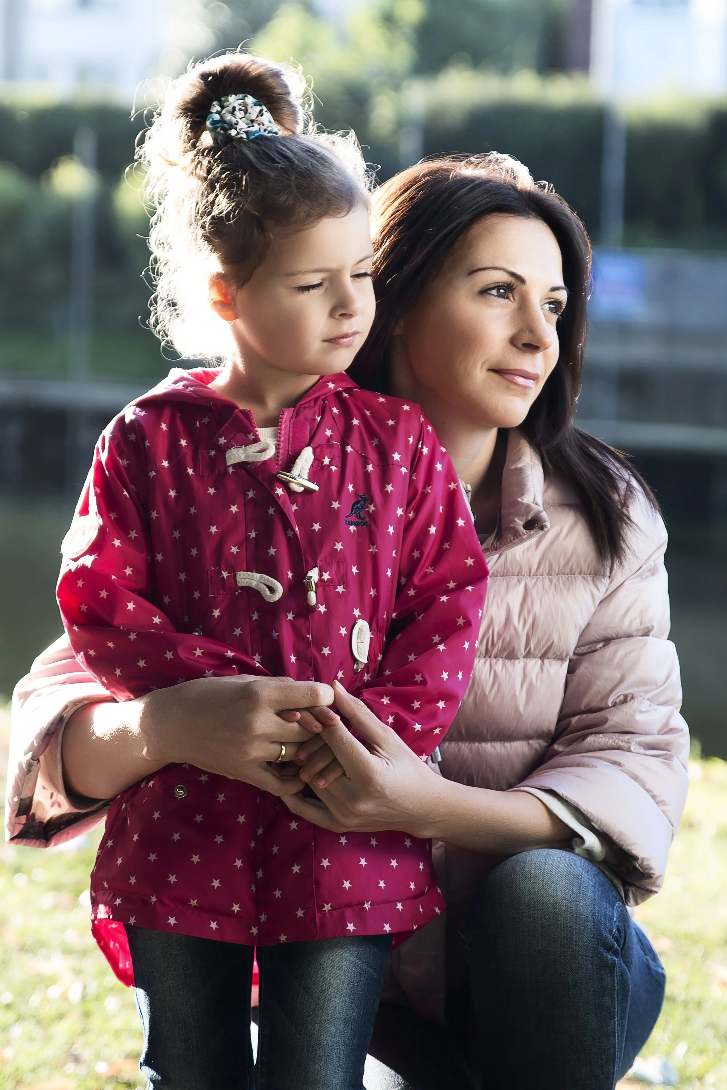Portrait of a woman and her daughter outdoors, bathed in warm sunlight. The woman, in a light pink jacket, gently holds the girl, who wears a red coat with stars, conveying a sense of warmth and connection.