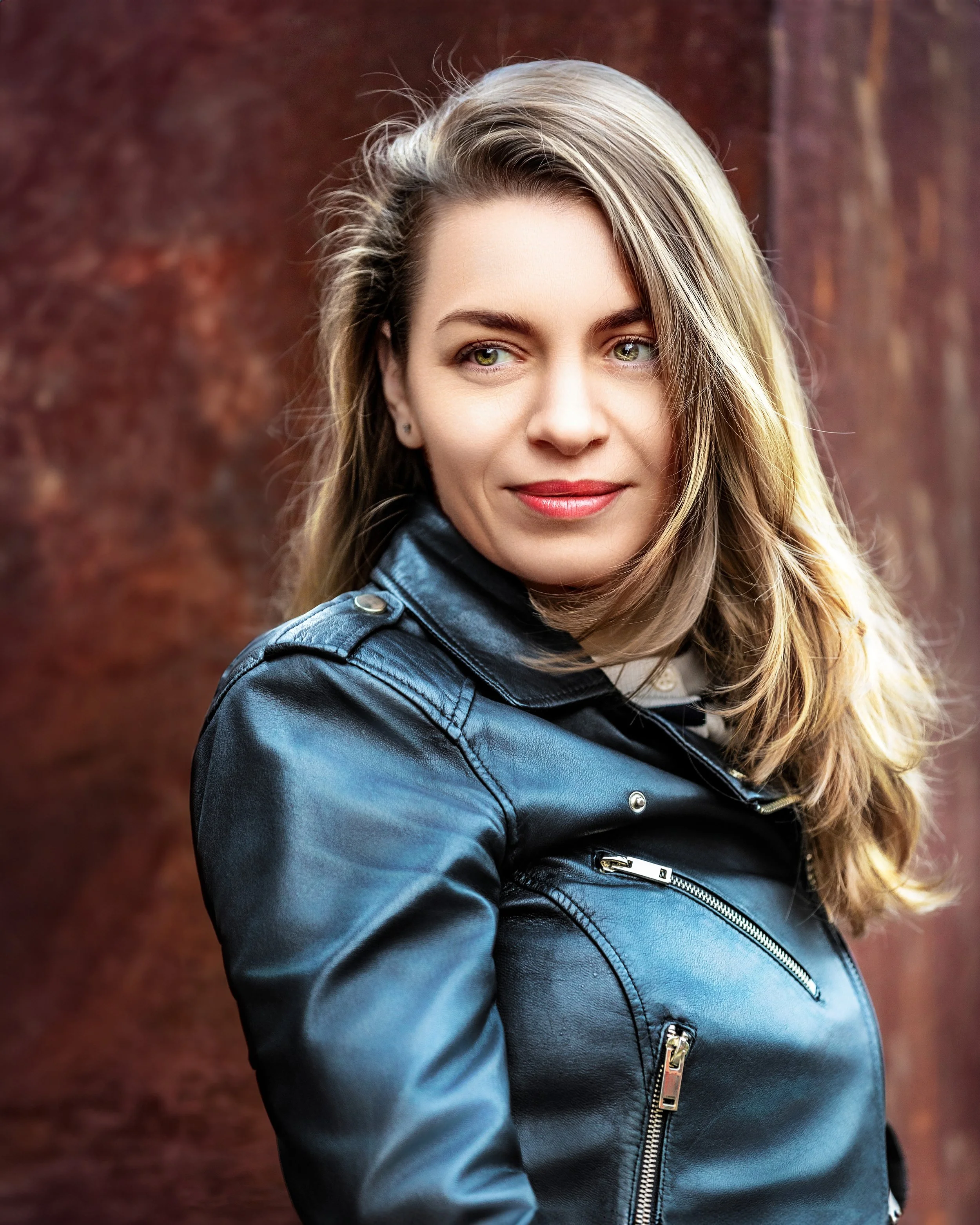 Portrait of woman with dark blonde hair in a leather jacket against a rusty building in Gloucester - England