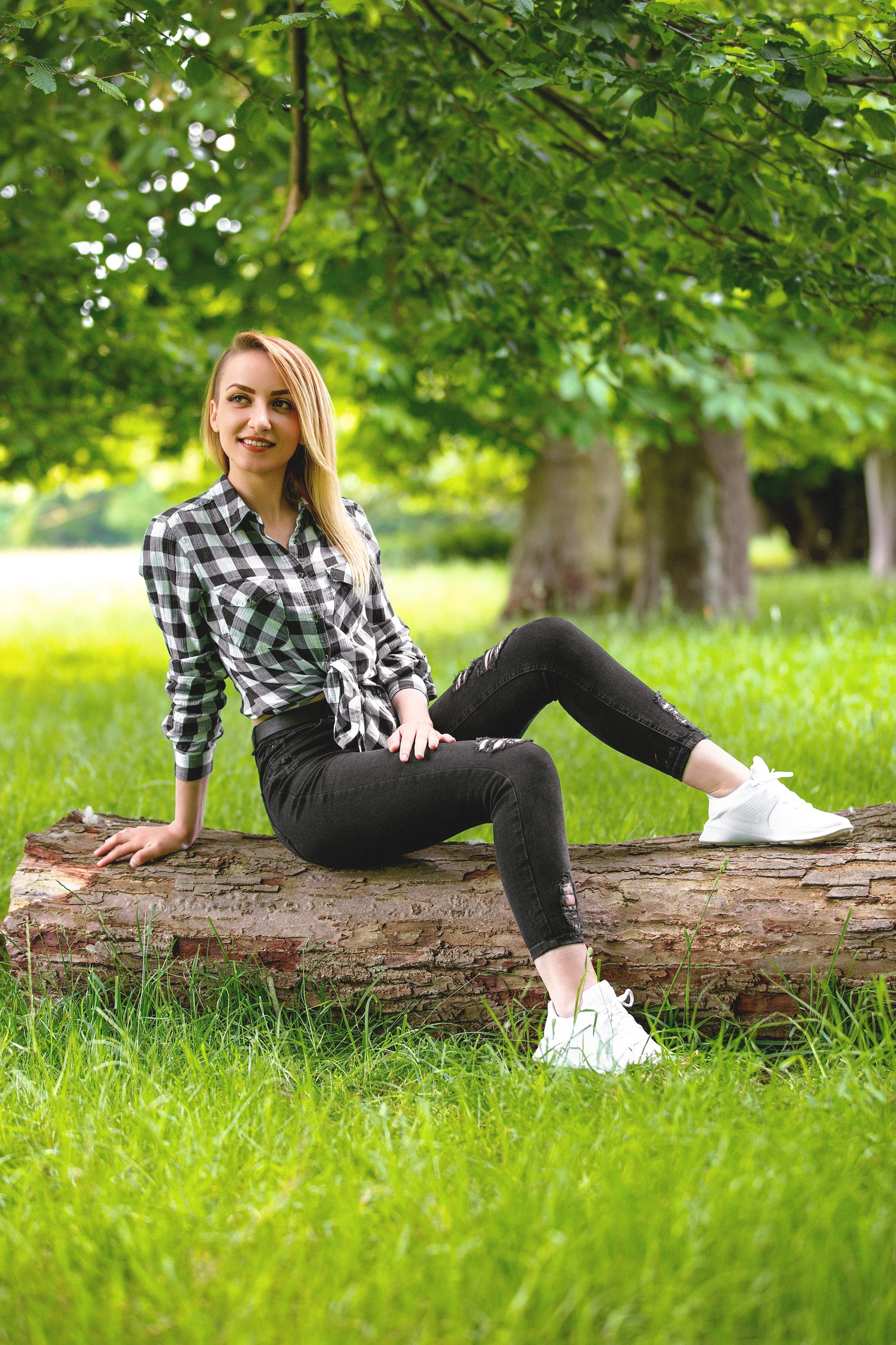Portrait of a woman sits on a log in a lush  green park, smiling and relaxed. She wears a black and white shirt and a black jeans. Trees fall out of focus in the background 