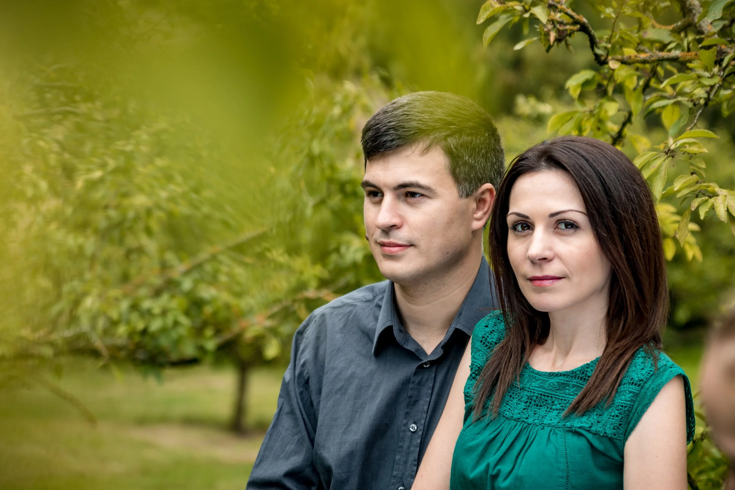 A man and woman sit closely in a lush garden. The woman wears a green dress, and the man a dark shirt. Both appear calm and content.