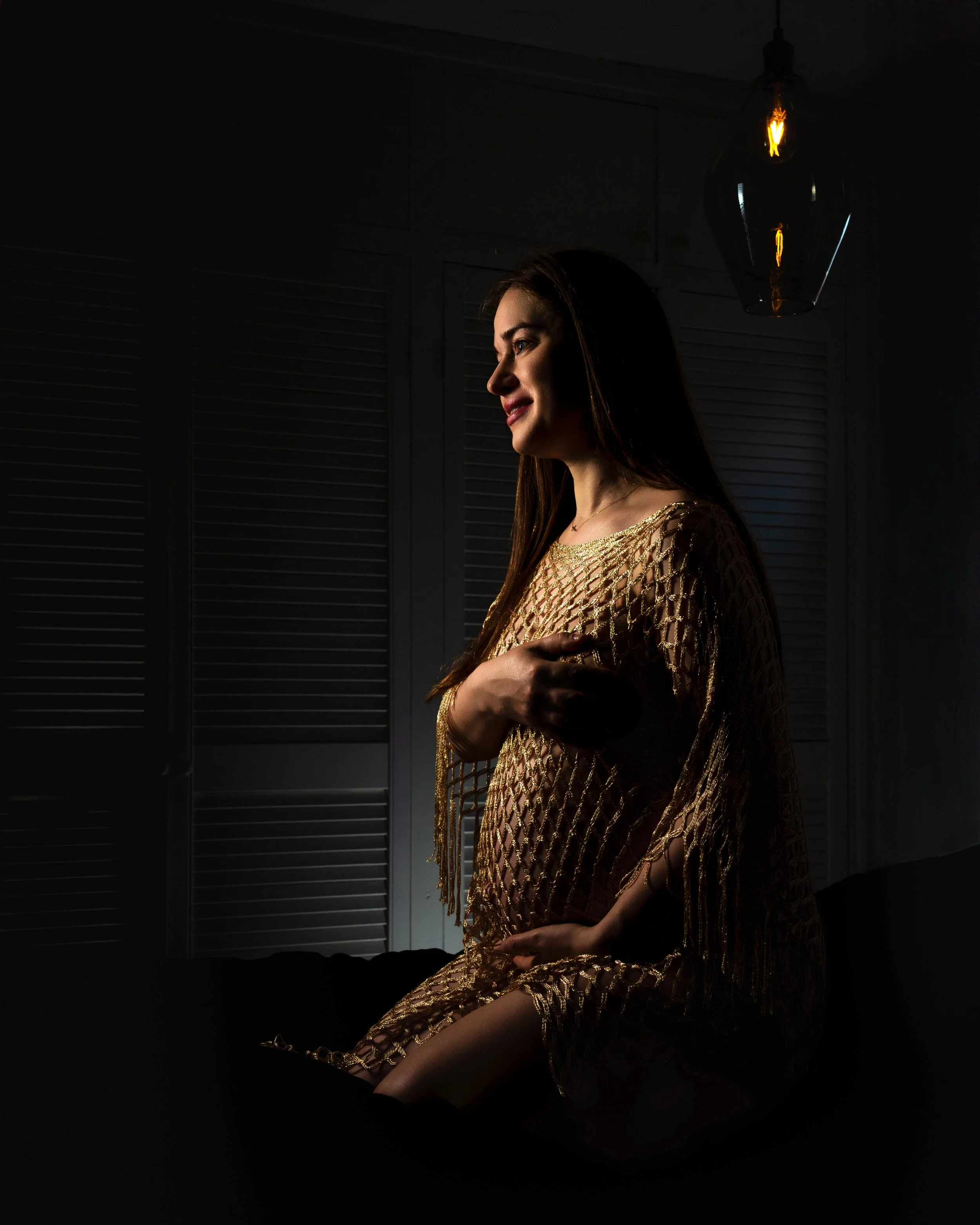 Pregnant woman sits in dim light, wearing a golden fishnet dress, gazing peacefully to the side. Warm, soft glow highlights her face and belly.