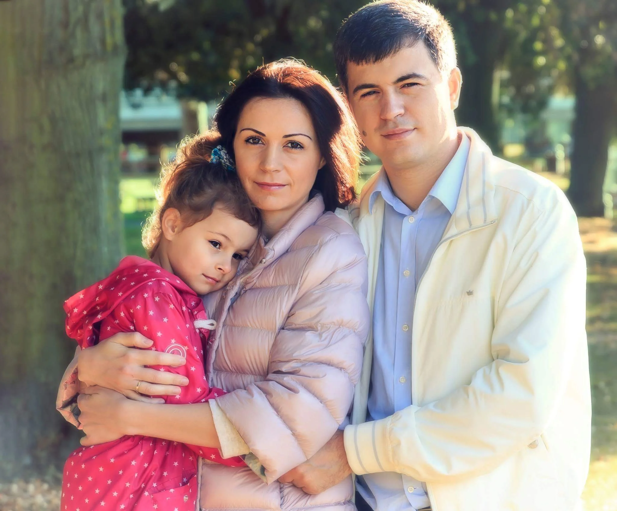 A family of three stands outdoors in a park, embraced warmly. The woman in the center holds a little girl in a pink coat with a slightly shy expression, while a man stands beside them, smiling gently. Sunlight filters through the trees, creating a se