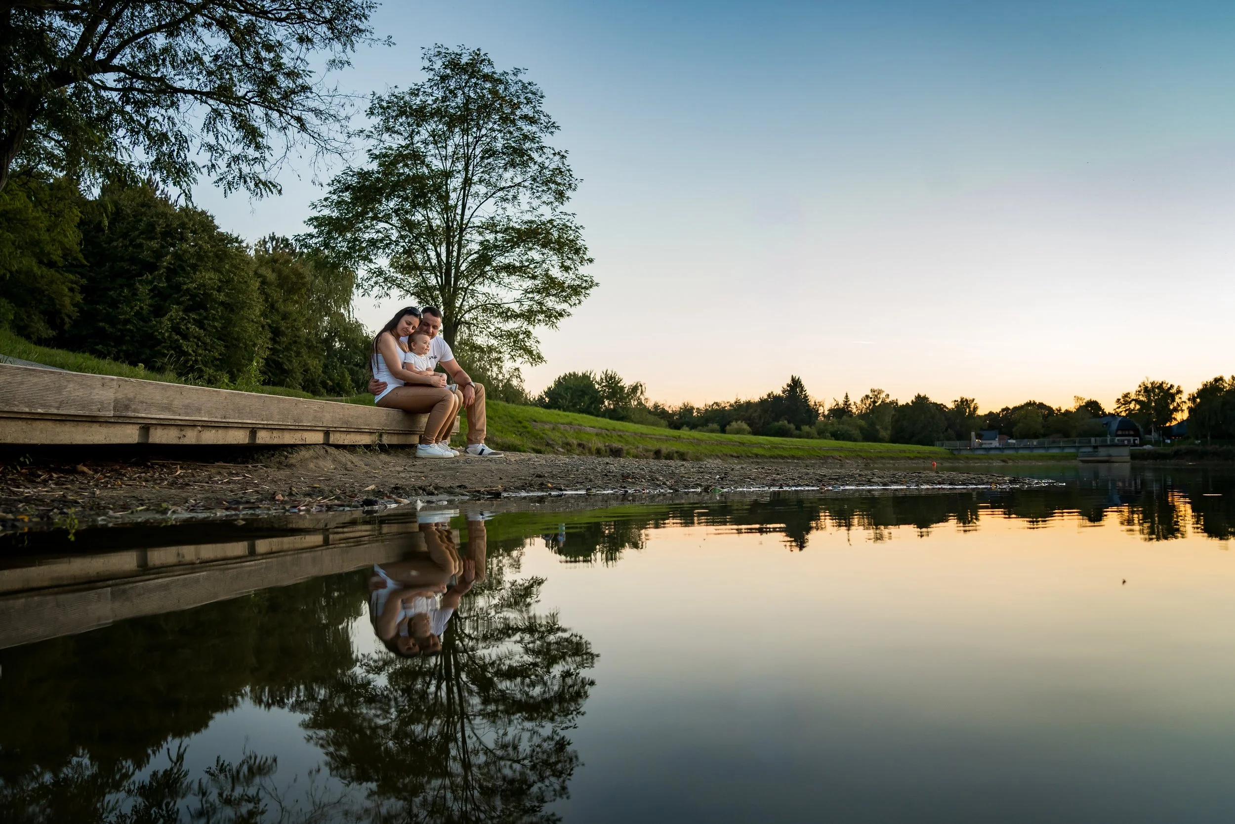 A couple and their baby sits on a wooden dock by a tranquil lake at sunset. Trees line the horizon, and their reflection shimmers on the calm water, creating a serene and intimate atmosphere.
