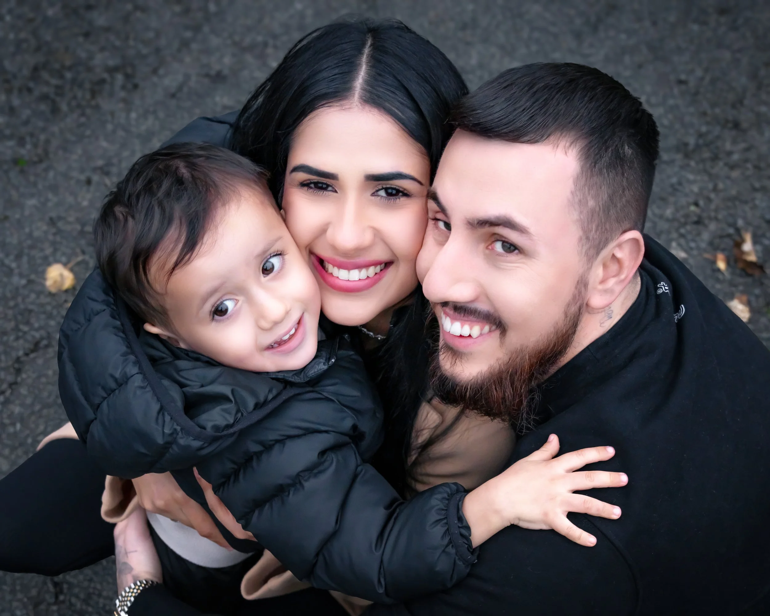 A joyful family portrait showing a man, woman, and child huddled together, all smiling warmly. The mood is affectionate and happy.