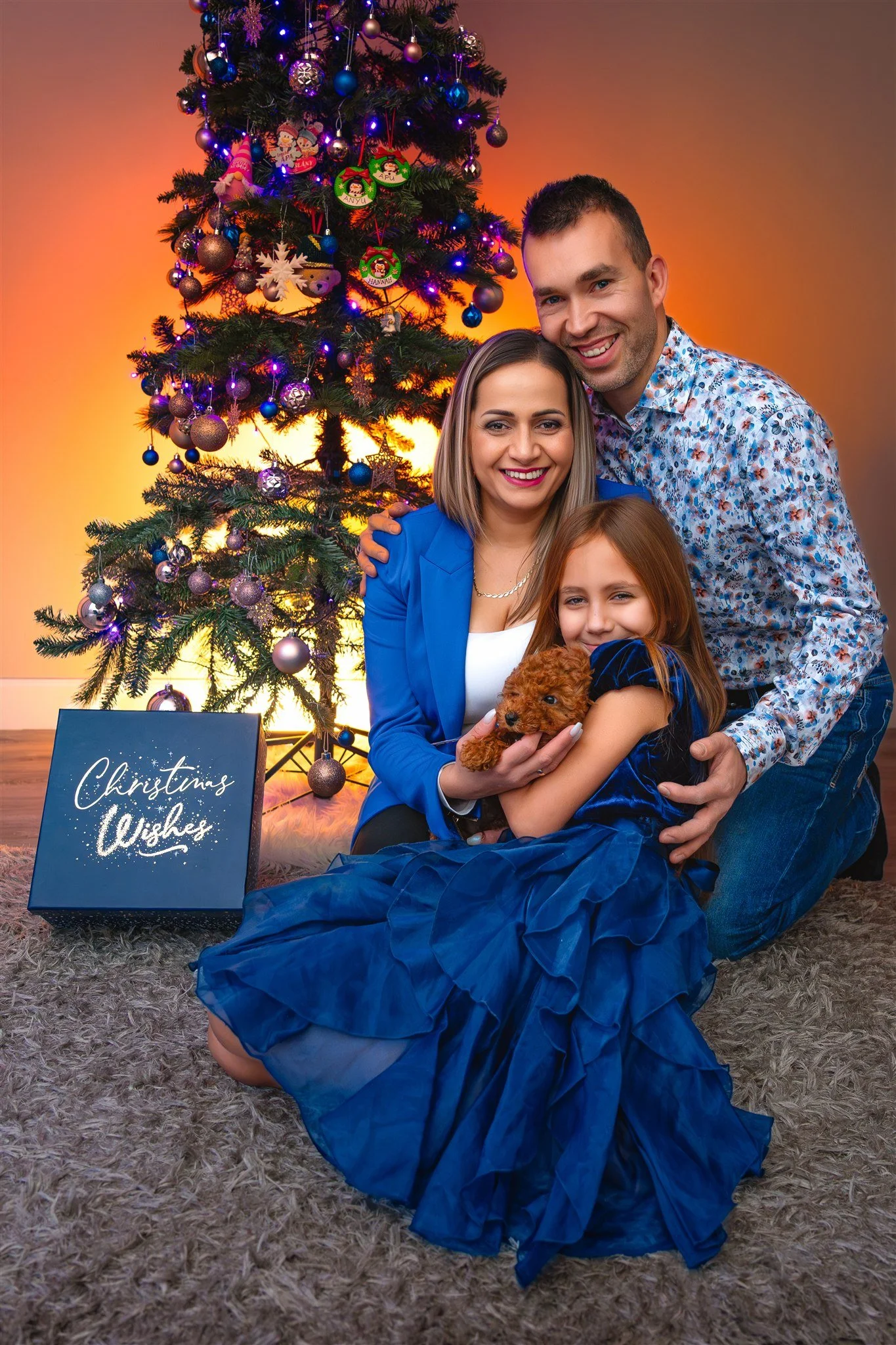 Portrait of a joyful family of three poses in front of a Christmas tree with blue and purple ornaments. The young girl holds a small brown dog. they sit beside a gift box which reads "Christmas Wishes". Warm, festive atmosphere.
