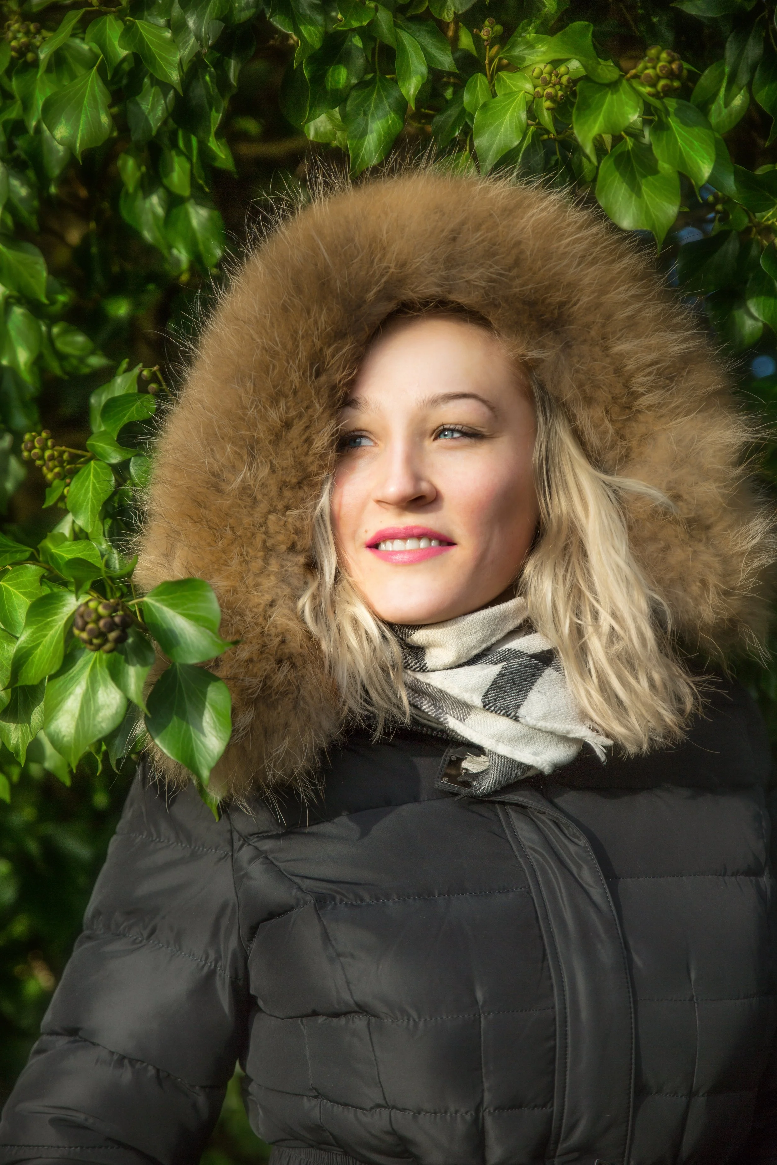 Portrait of a blonde woman in black winter jacket with a fury hoodie on her head. She stands against a lush green bush