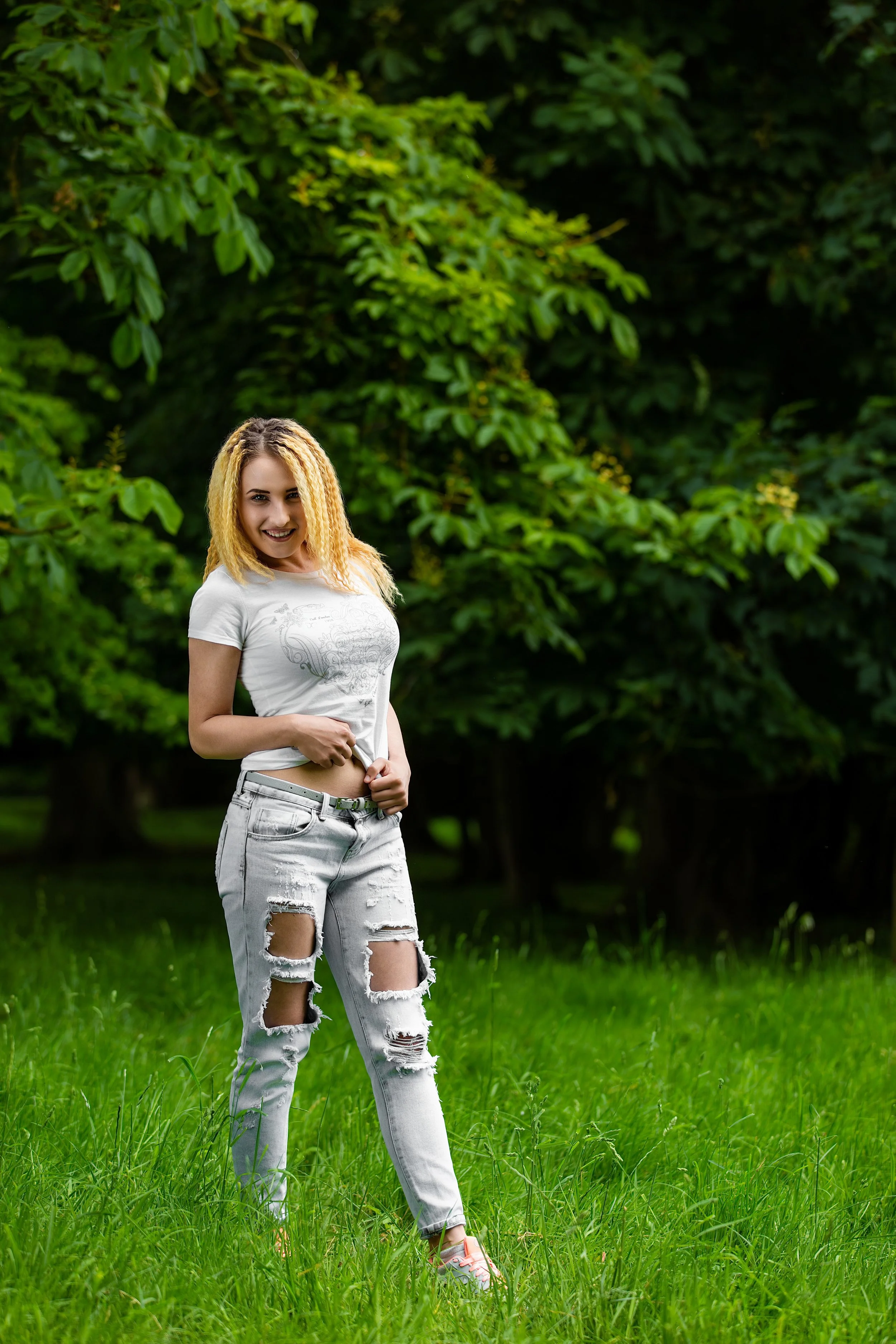 Portrait of a young blonde girl wearing white T-shirt and ripped jeans on a field by the forest in the spring