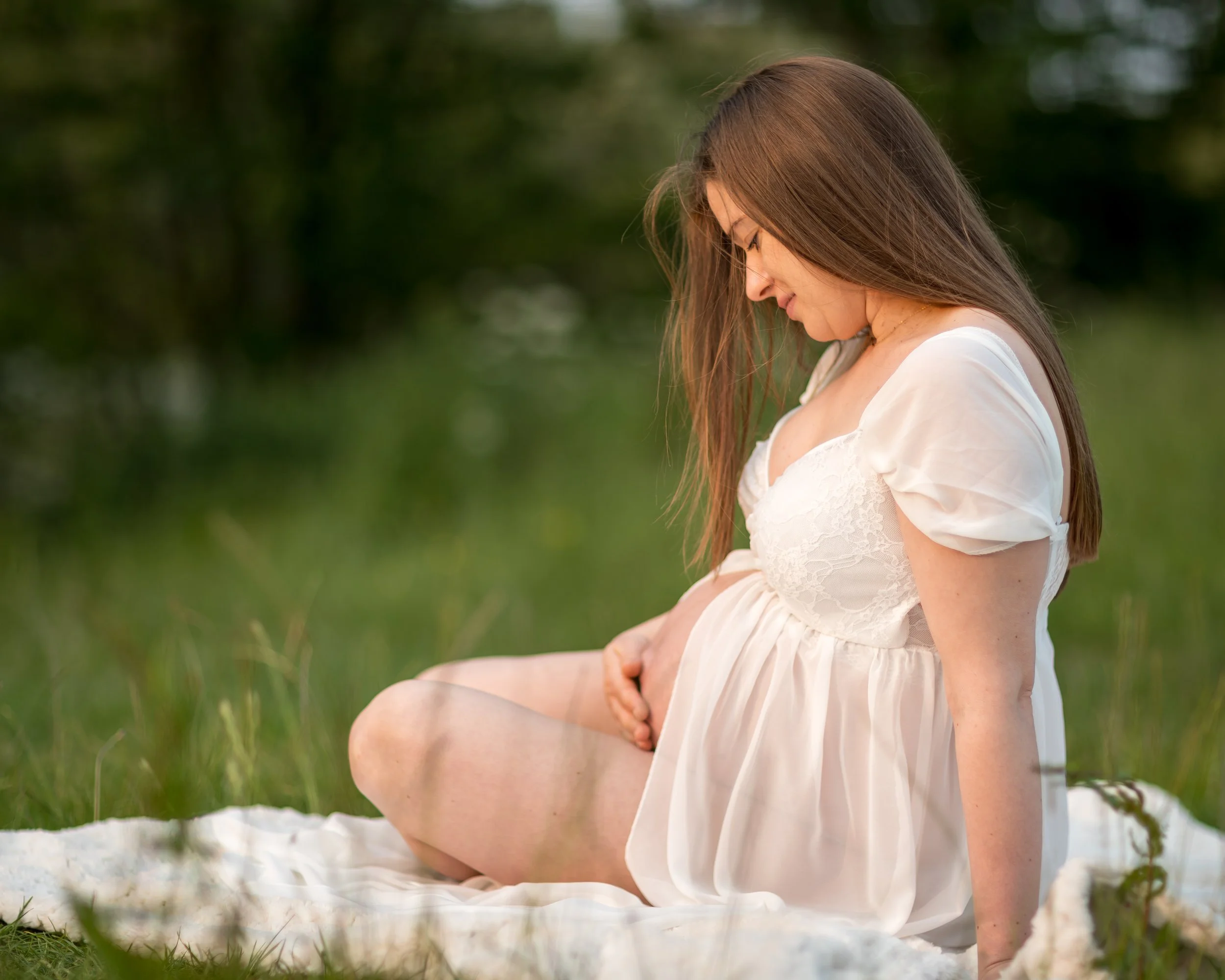 Pregnant woman in a white dress sits on a blanket in a grassy field, gently cradling her belly. The atmosphere is serene and focused.