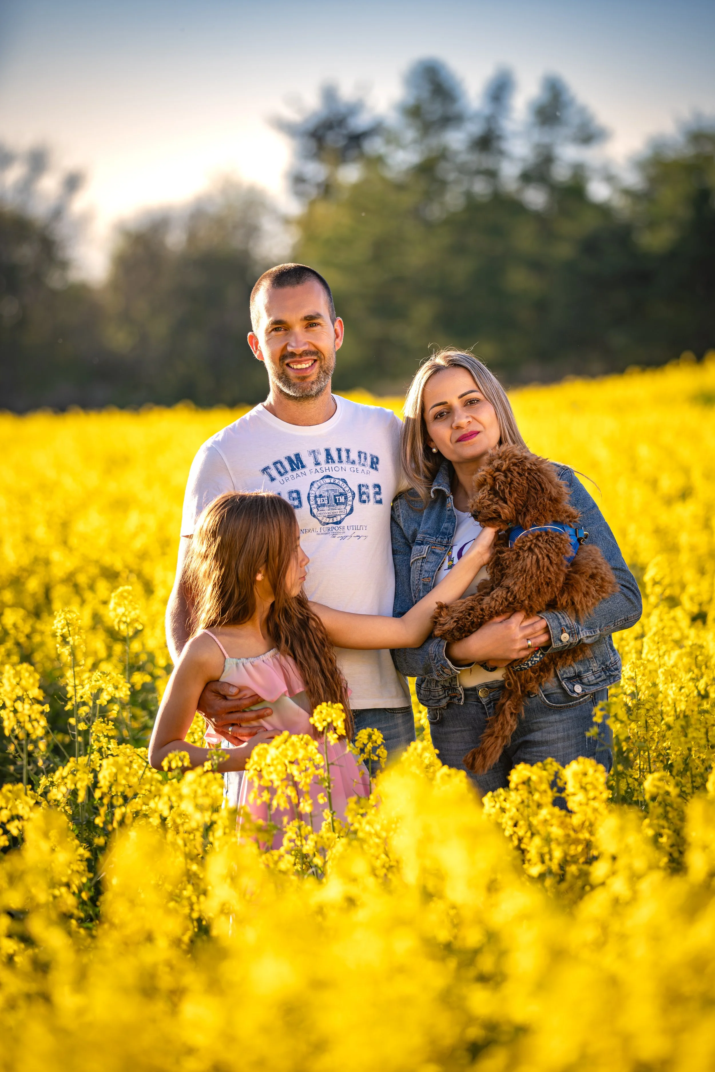 A family enjoying a warm spring evening in a blooming rapeseed field, gathered together with their small dog in soft golden sunlight.