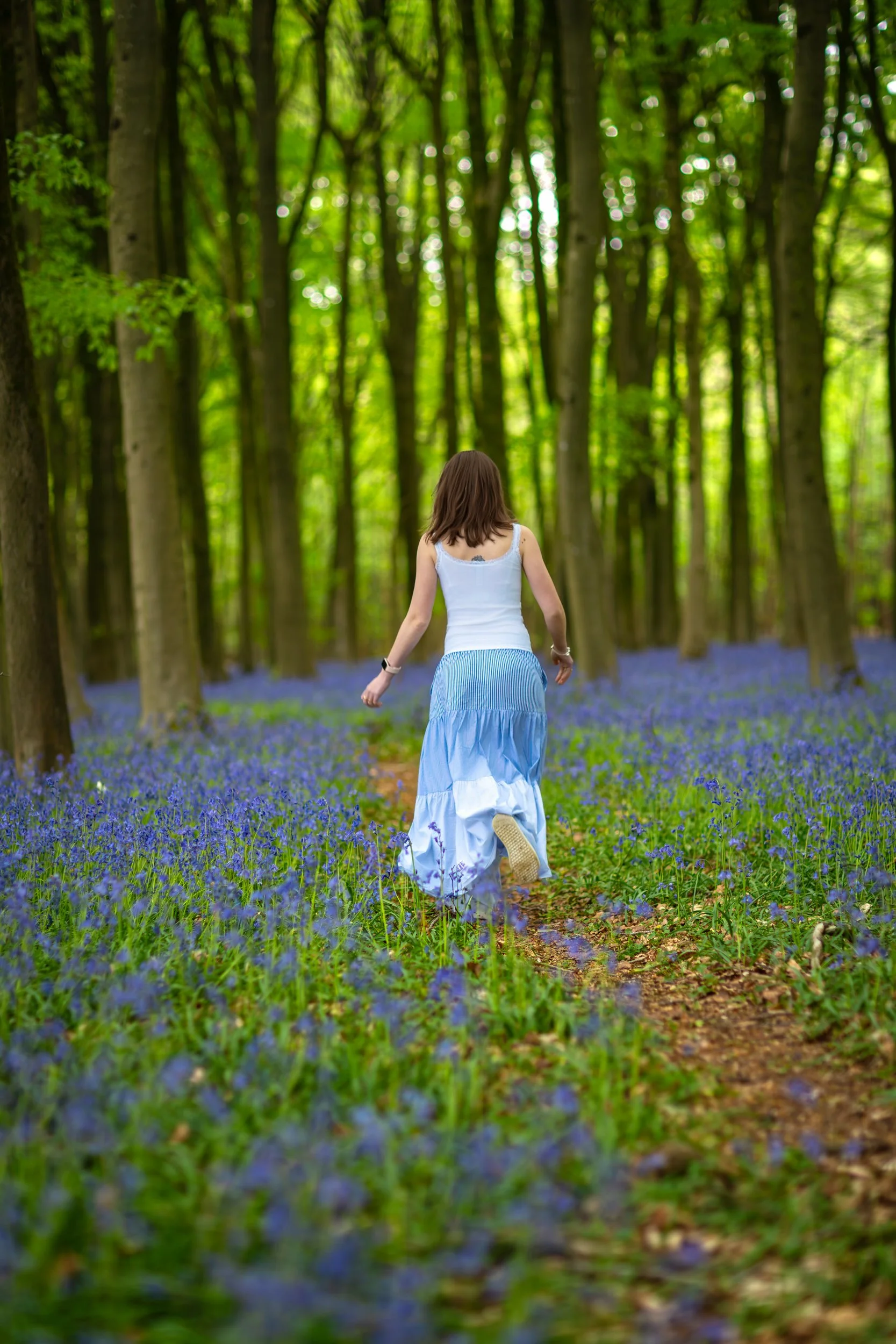 A young girl walking along a woodland path surrounded by blooming bluebells, with soft spring light filtering through the trees.
