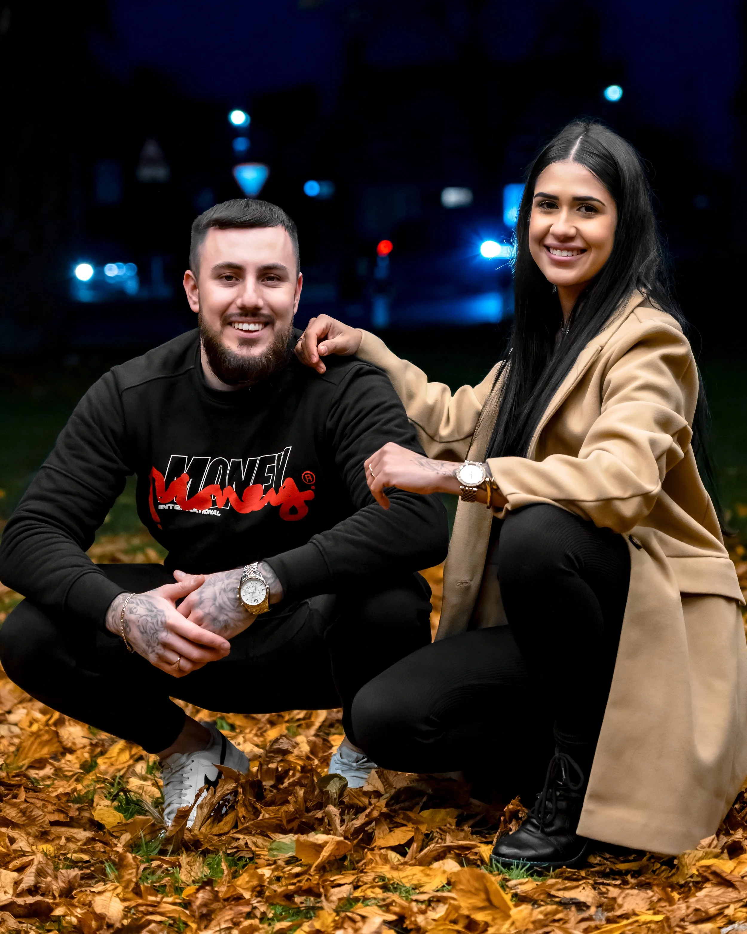 A smiling man and woman pose outdoors at night, surrounded by fall leaves. He crouches in black attire; she kneels in a tan coat, exuding warmth and joy.
