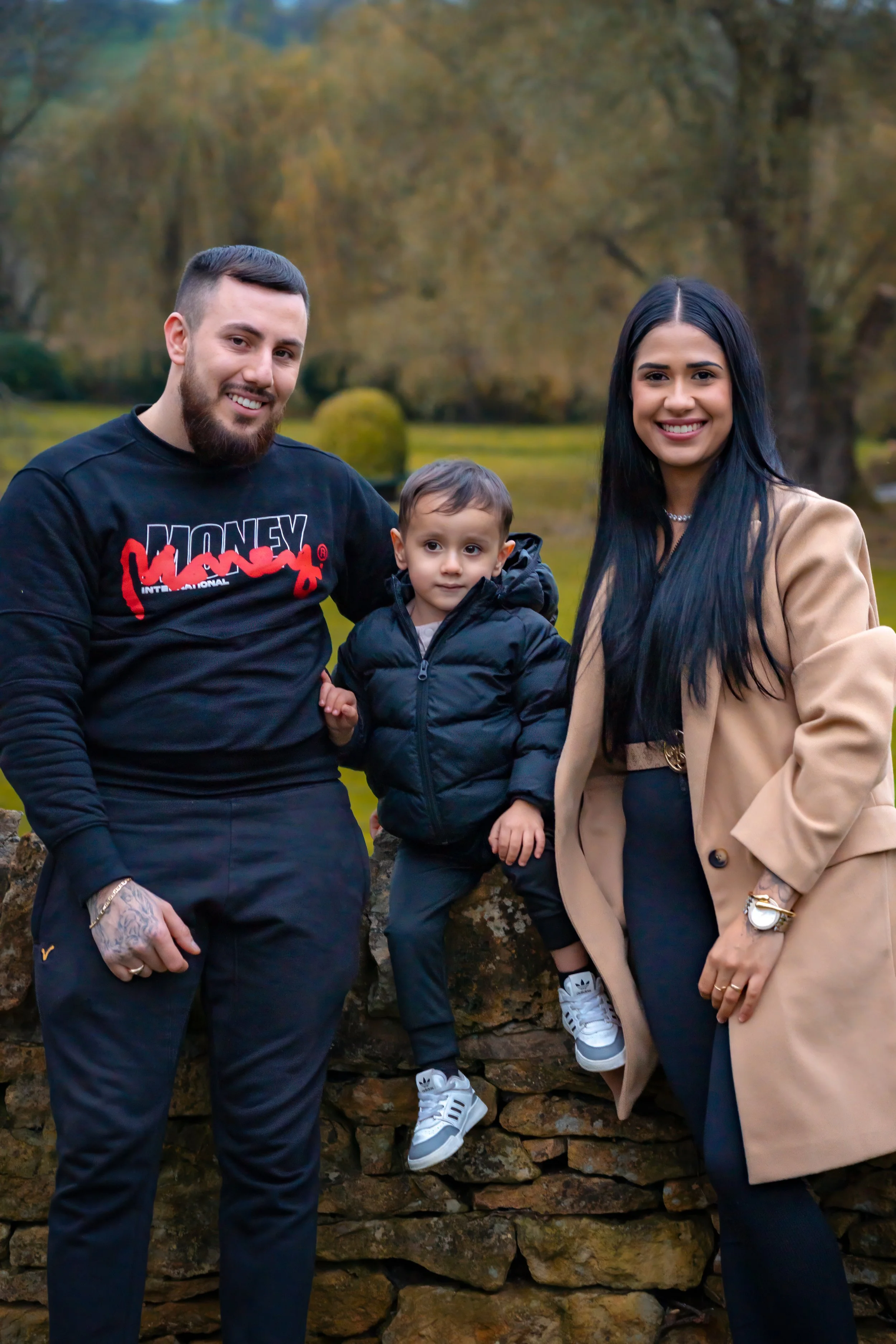 A smiling family stands outdoors by a stone wall. A man in a black sweater and a woman in a tan coat flank a little boy in a black jacket. The setting is a park with autumnal trees, conveying a warm, joyful atmosphere.