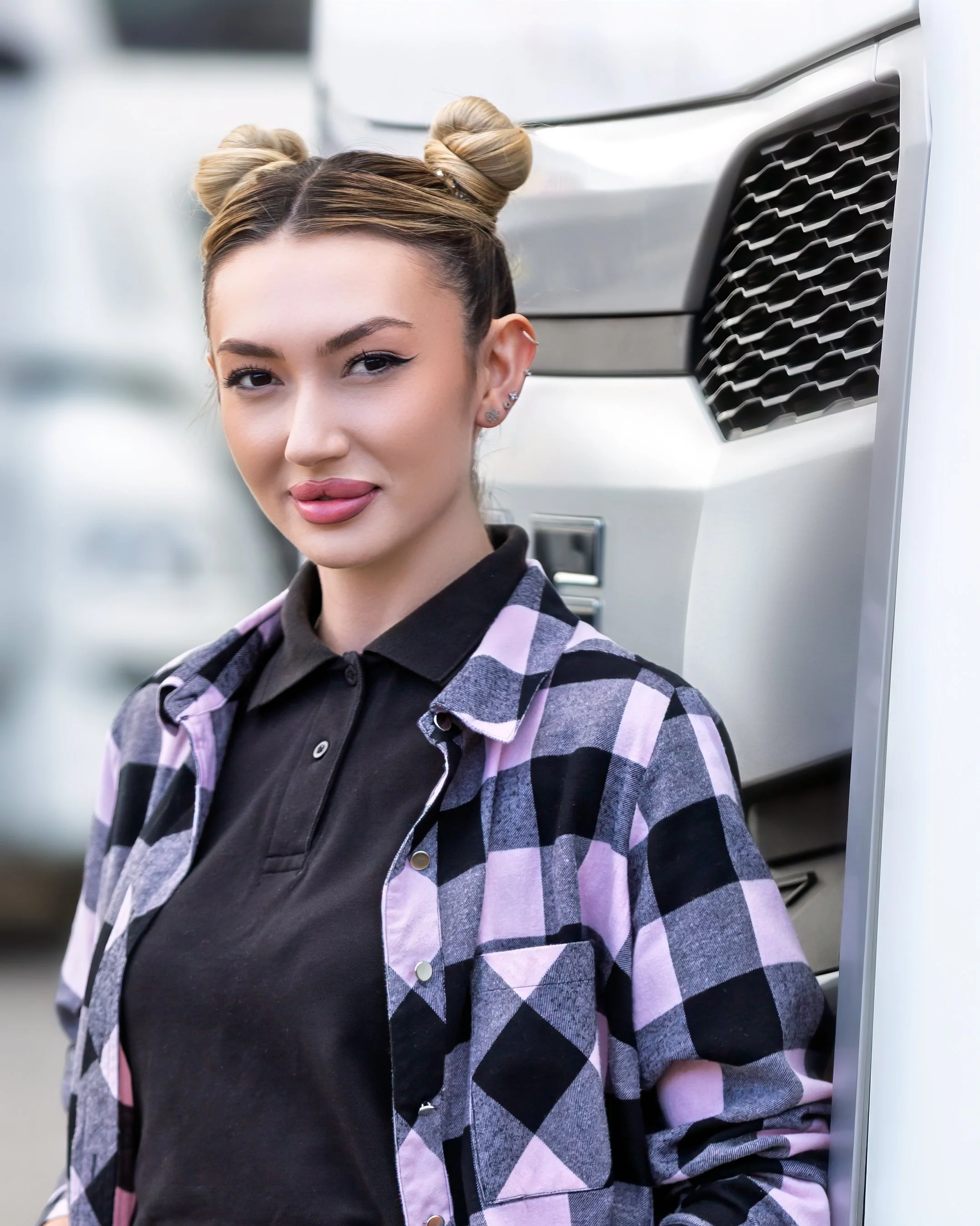 Portrait of a young woman in front of a large white DAF lorry. They wear a pink checked shirt