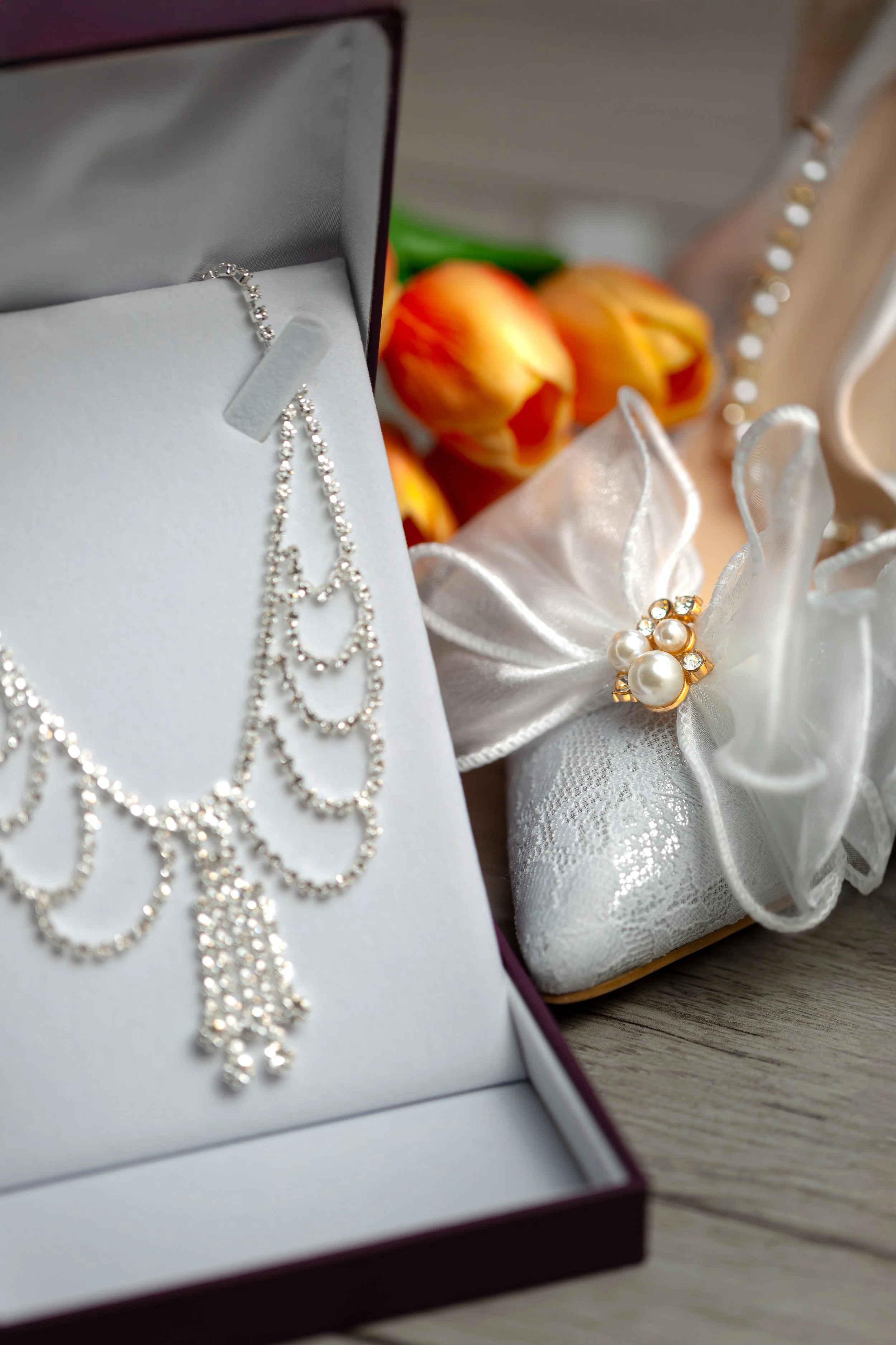 A delicate silver necklace with cascading chains in an open box beside white high heels adorned with pearls and bows, with orange flowers in the background.