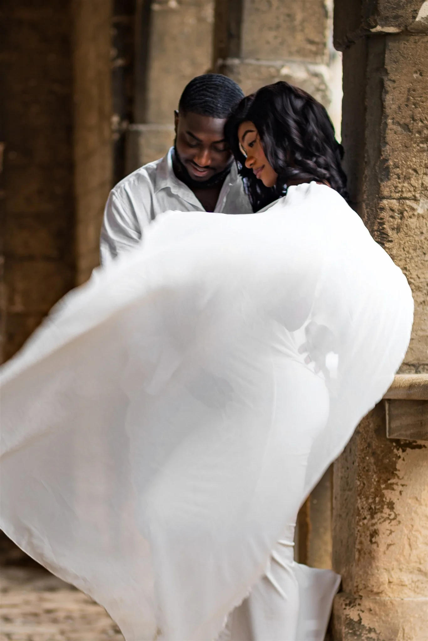 A couple stands in an embrace, with the pregnant woman's flowing white dress billowing dramatically. They smile tenderly amid ancient stone architecture, conveying romance.