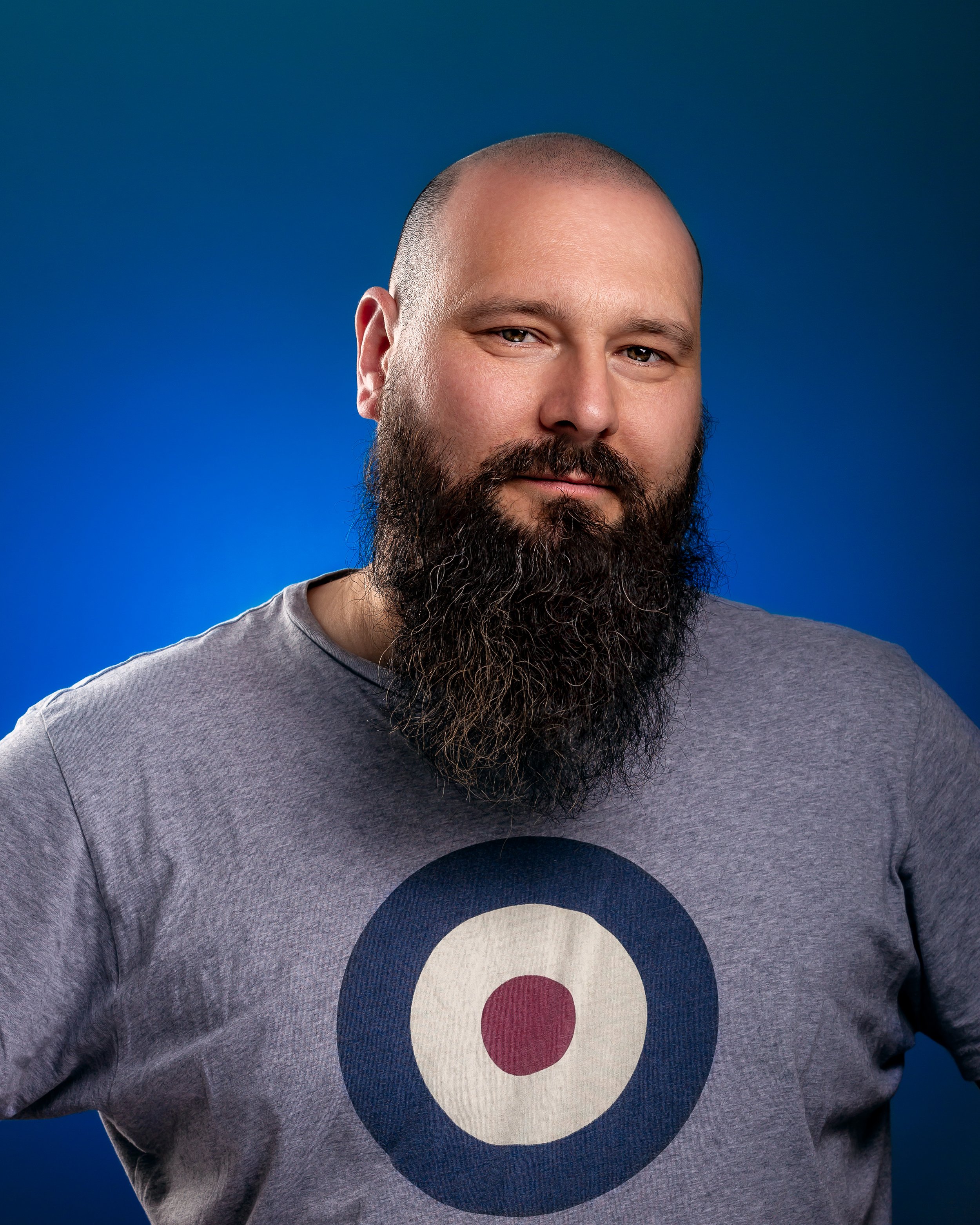 Bearded man in a great t-shirt with a mod target design, standing against the gradient blue background. He is relaxed and confident.