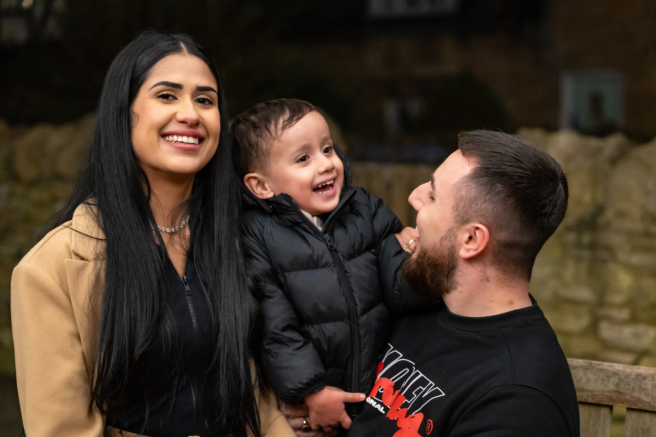 A joyful family moment: a woman in a beige coat and a man in a black shirt, holding a smiling toddler in a puffy jacket, all sharing laughter outdoors.