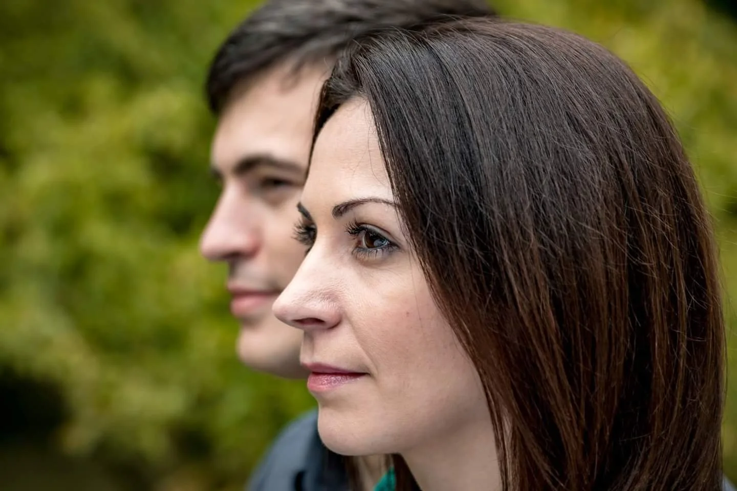 Close-up of a woman in focus, gazing thoughtfully to the right with a slight smile. A man in the background is blurred. Greenery is visible behind them.