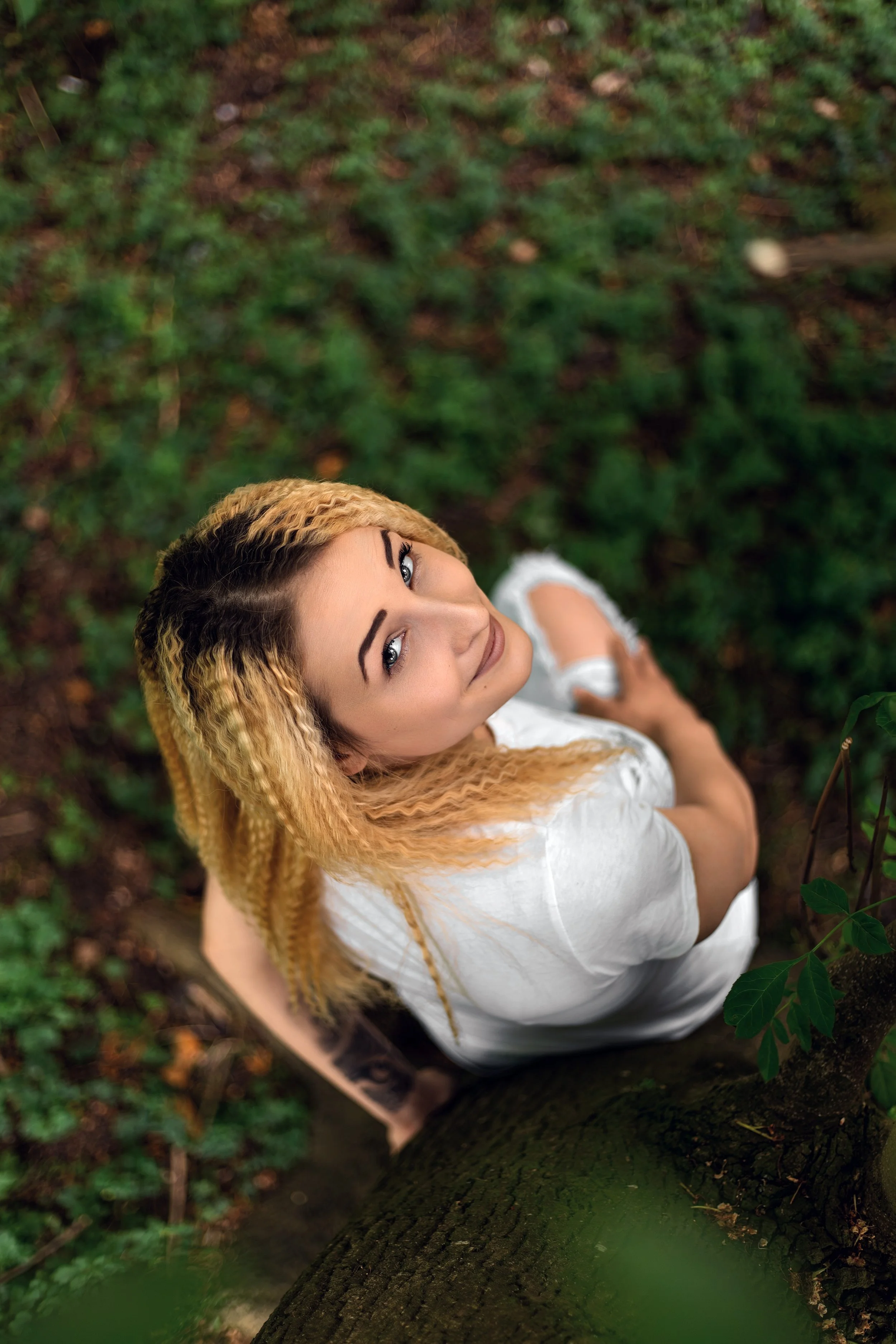 Portrait of a woman with blonde hair looking up at the camera while sitting at the bottom of a tree. She wears a white T-shirt, ripped jeans,  surrounded by lush green foliage, conveying and serene contemplative mood