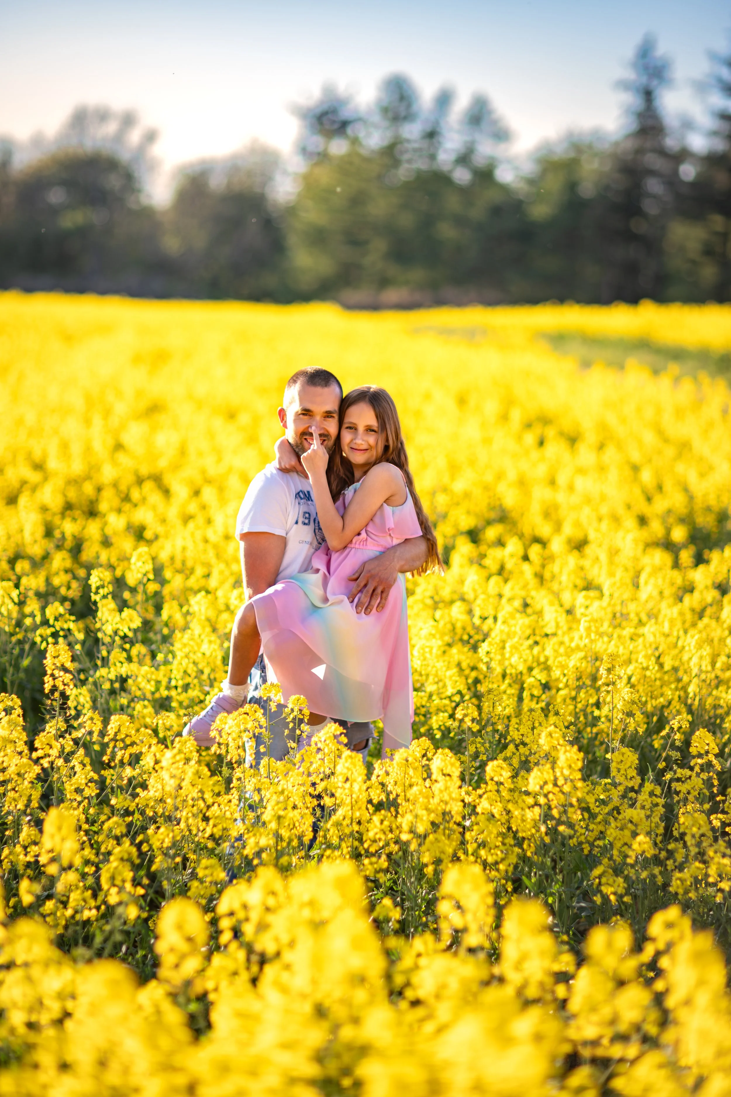A smiling father holding his daughter in a blooming yellow rapeseed field during golden hour, surrounded by warm sunlight and soft spring colors.