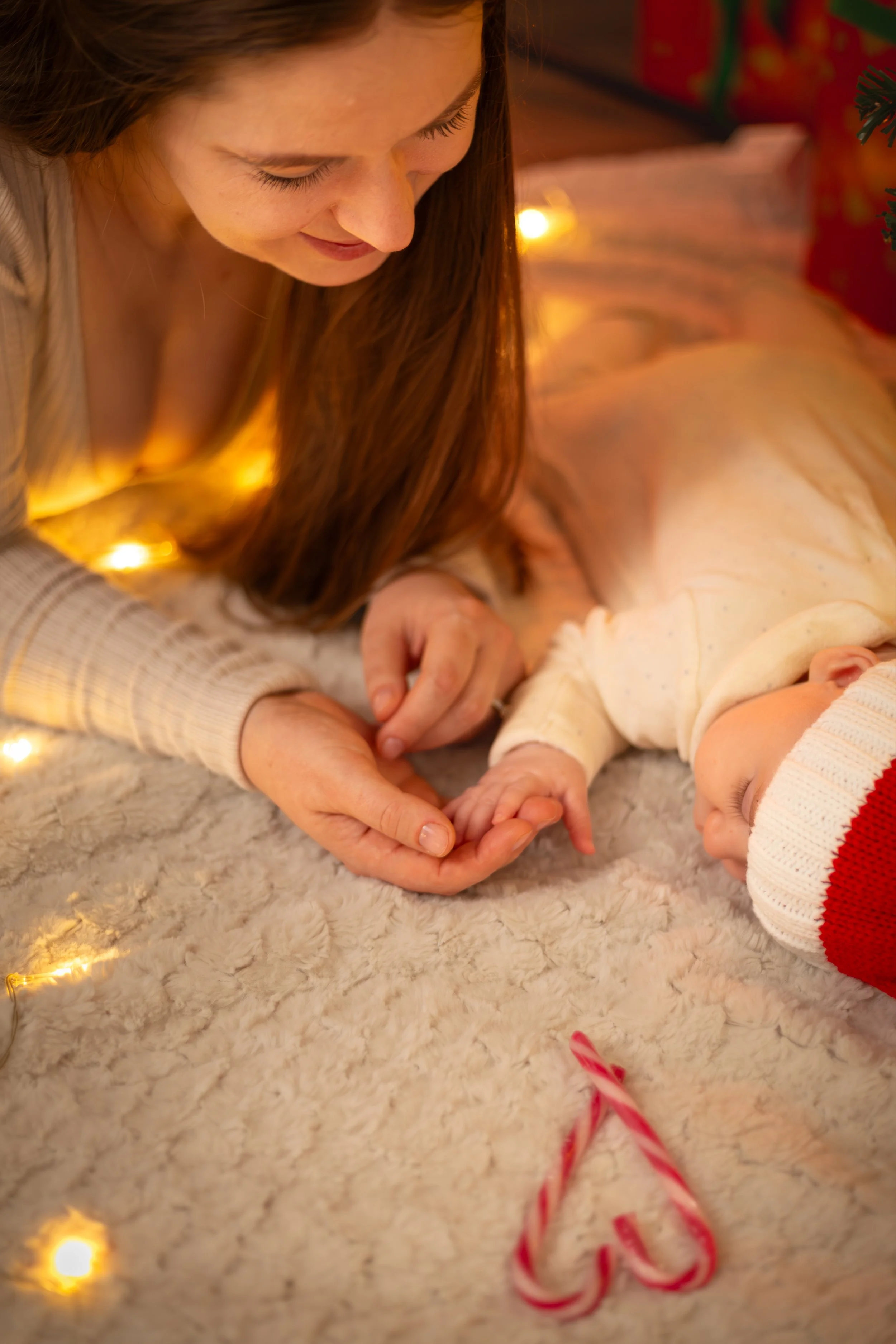 A woman lovingly holds a baby's hand on a cozy blanket, with warm string lights and candy canes forming a heart shape, creating a festive, tender atmosphere.