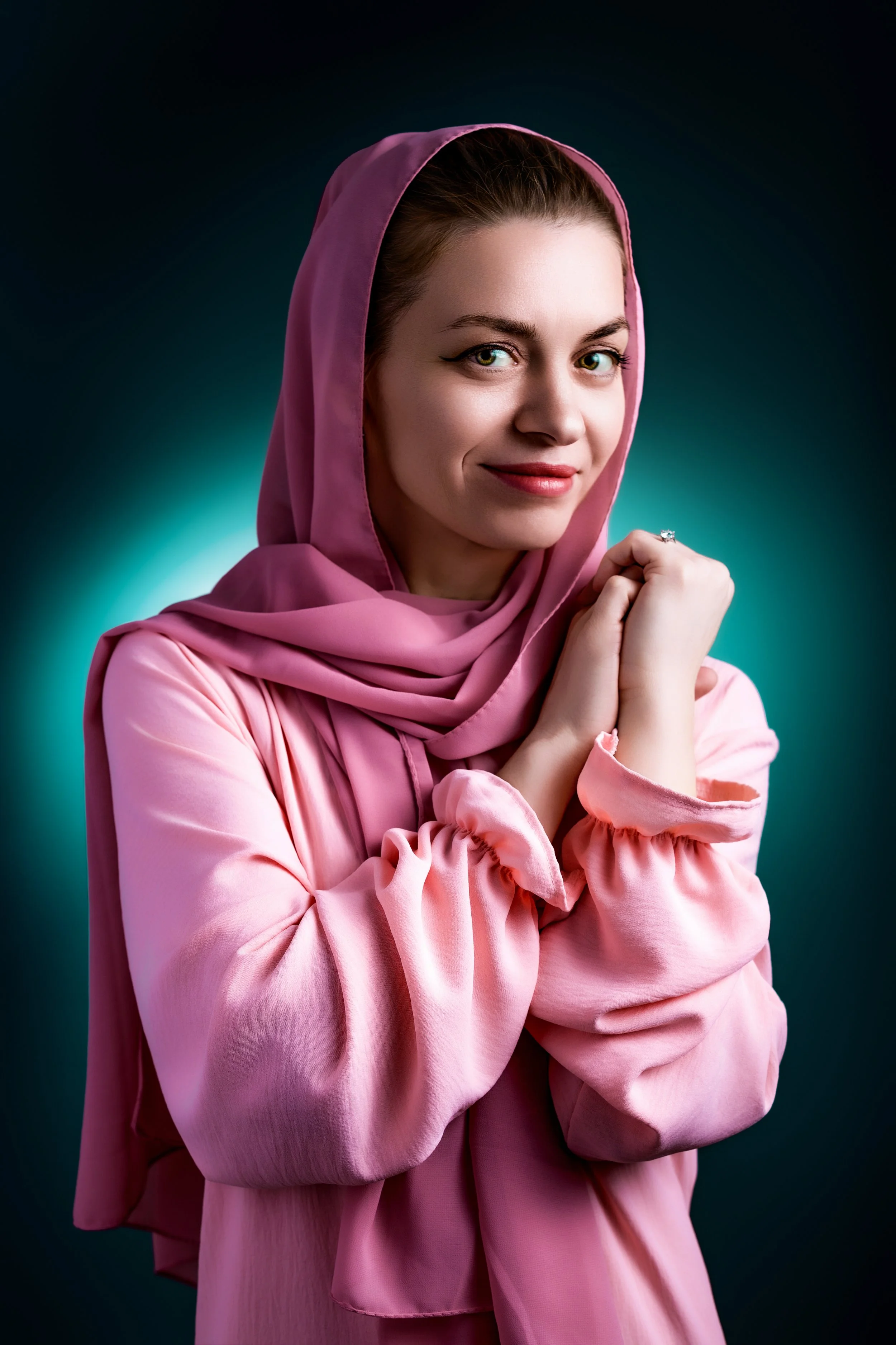 Studio portrait of a confident woman in a pink hijab and blues stands against a teal background, smiling softly with hands clasped. The mood is warm and welcoming