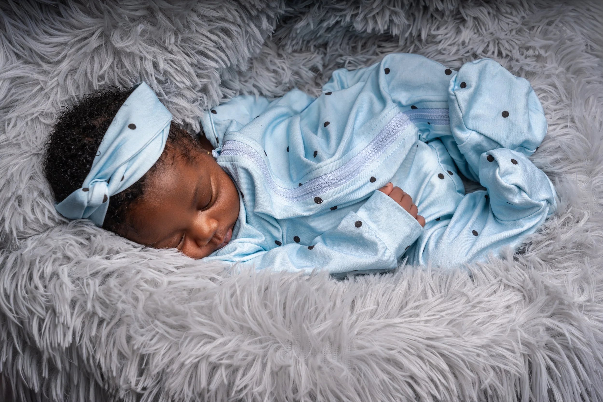 A sleeping baby lies on a fluffy, gray blanket, wearing a light blue outfit with black polka dots and a matching headband, conveying peace and warmth.